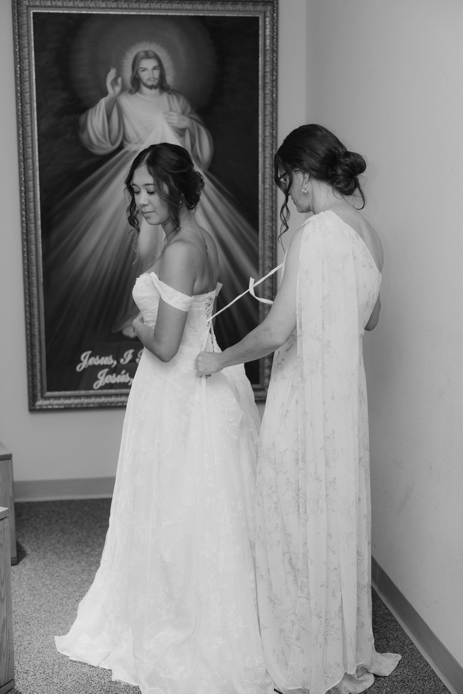 Black and white photo of bride getting wedding dress laced up before ceremony with Divine Mercy painting backdrop.