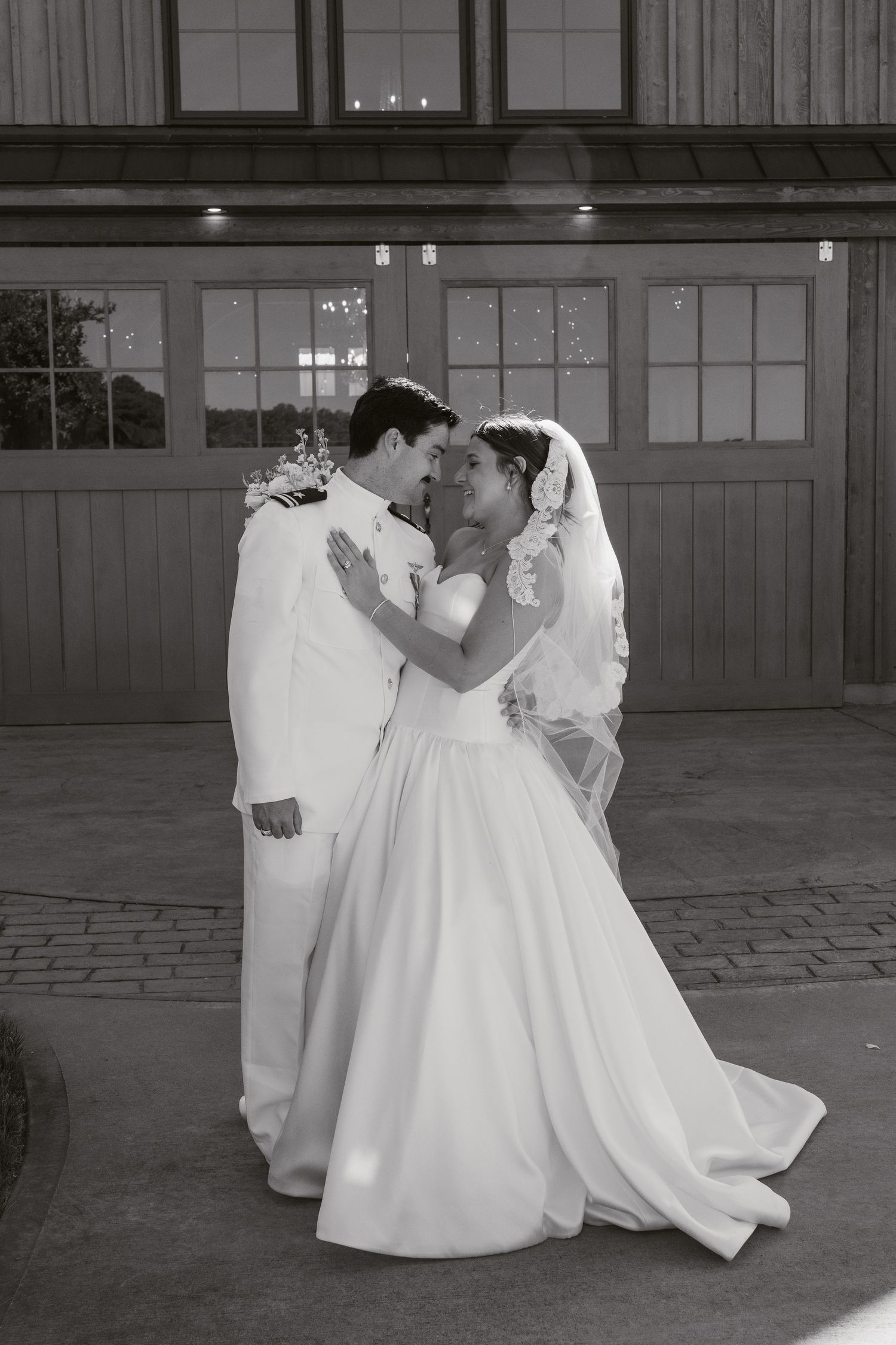 Black and white photo of a bride and groom sharing a romantic moment in wedding attire outside a venue.