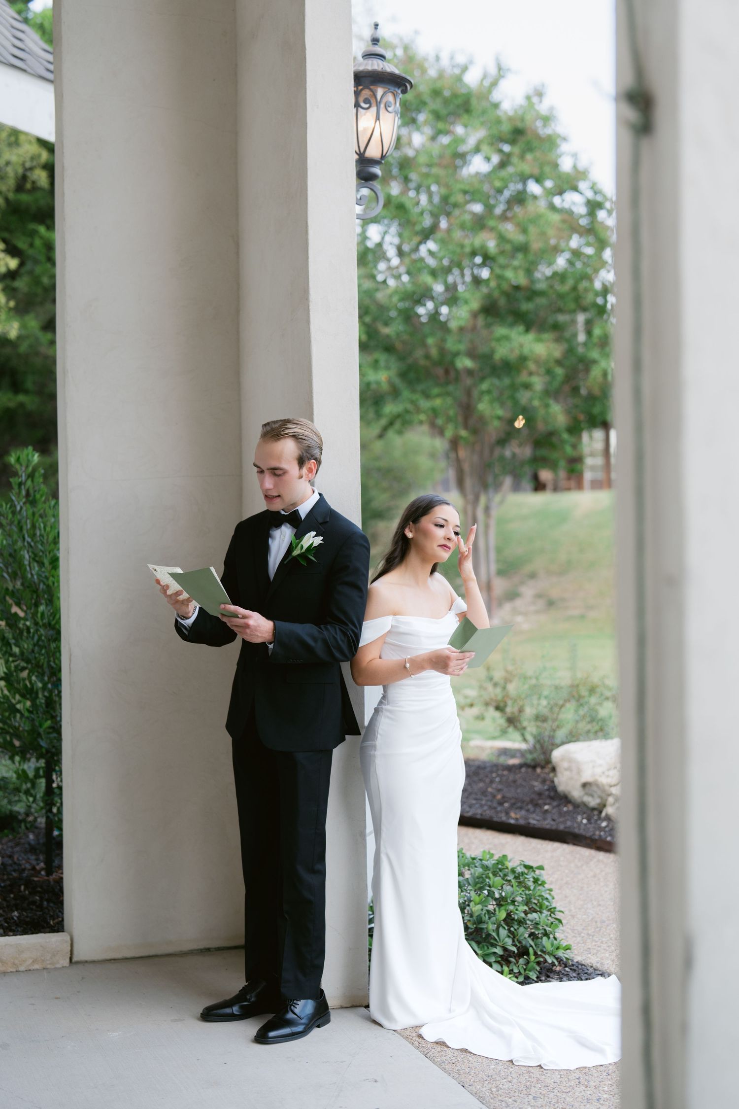 Bride and groom stand back-to-back reading vows beside a column, she in white gown, he in black tuxedo.