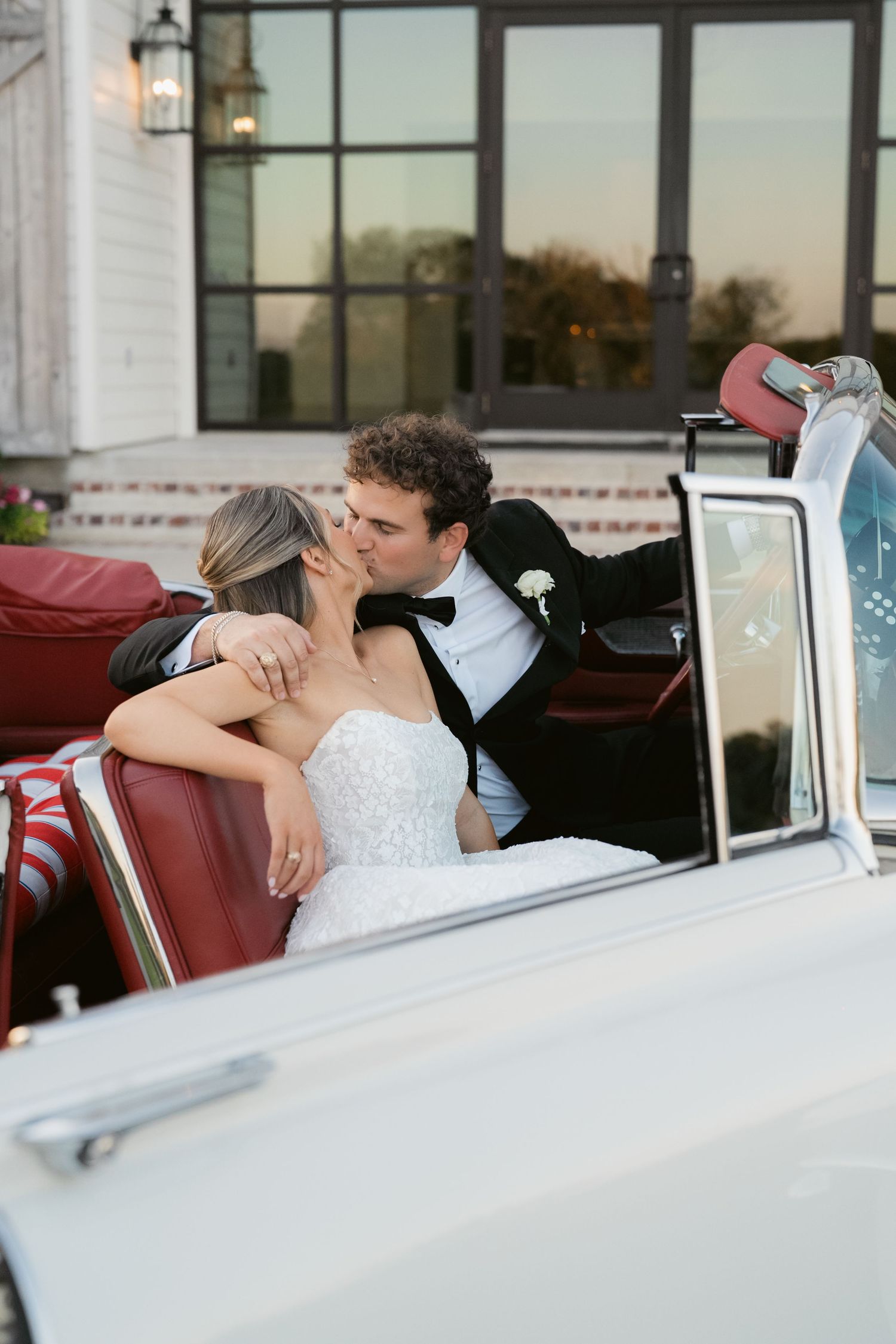 A bride and groom share a romantic kiss in a classic white convertible car with red interior on their wedding day.