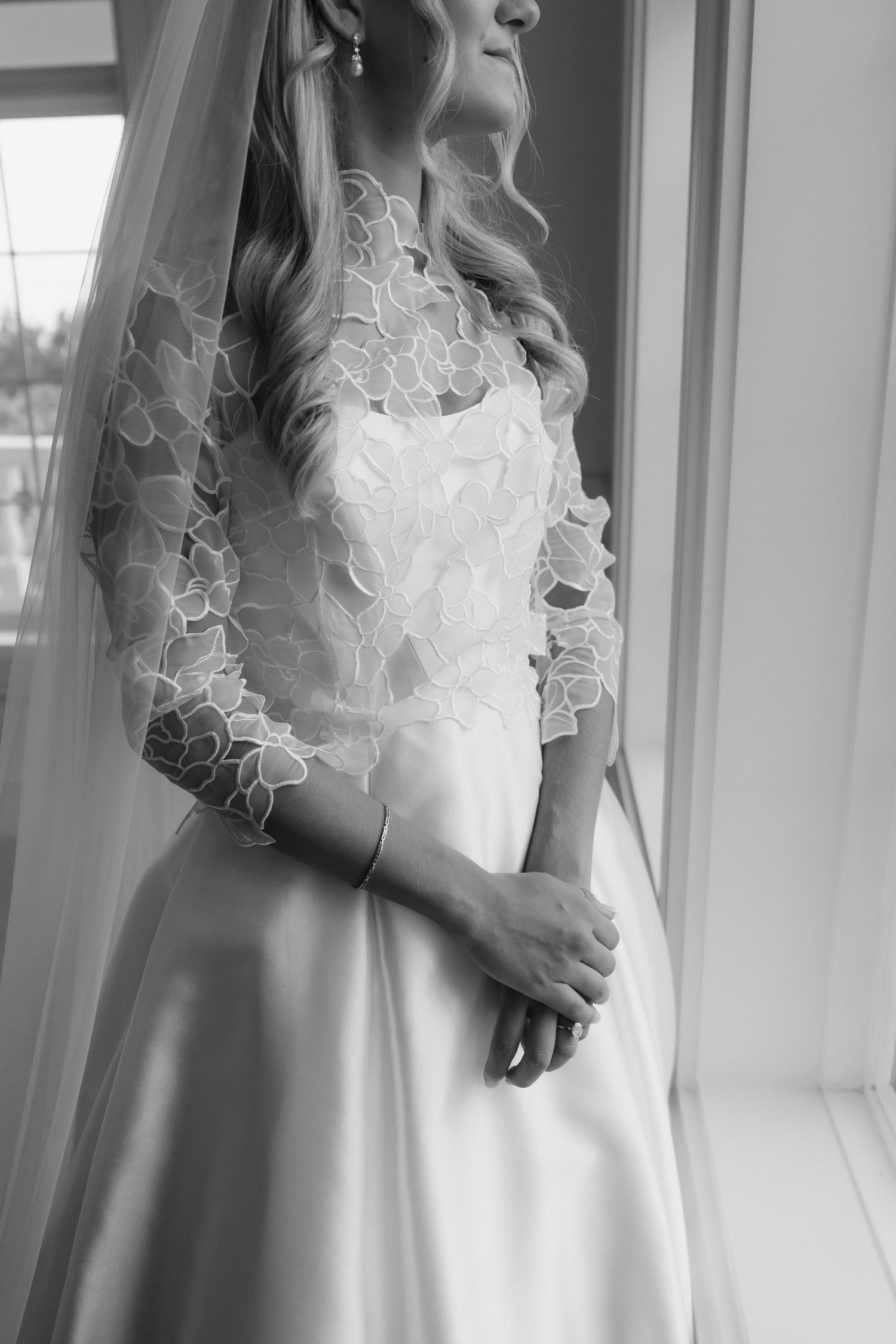 Black and white photo of a bride in lace wedding gown and veil standing by a window.