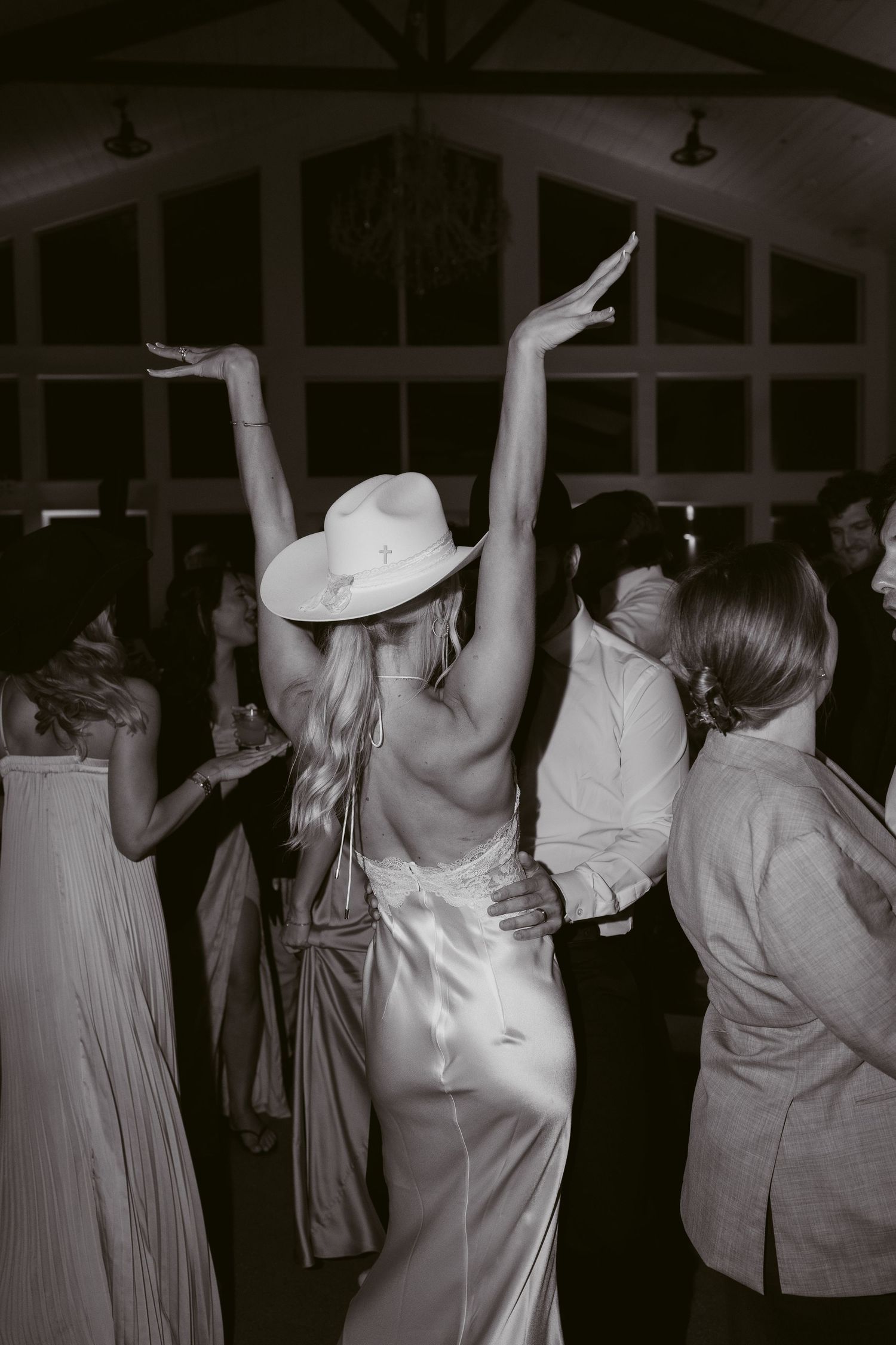 Bride in white satin gown and cowboy hat dances with arms raised at wedding reception.