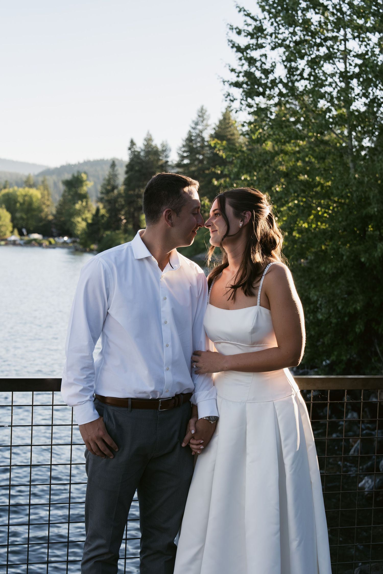 A couple in wedding attire share a romantic moment on a lakeside bridge surrounded by lush green trees.