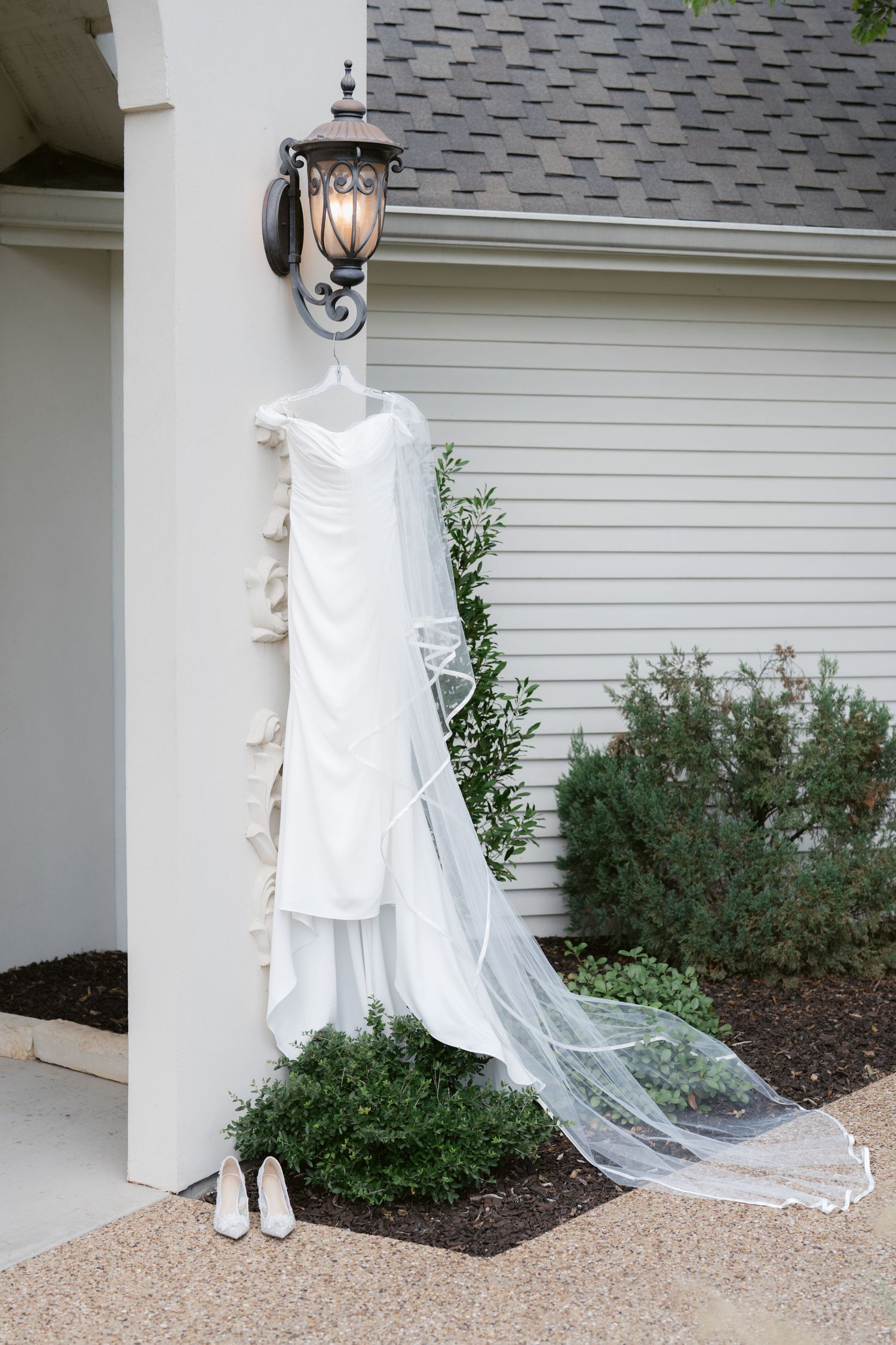 White wedding dress with long veil hanging on outdoor column beside bridal heels on patio.