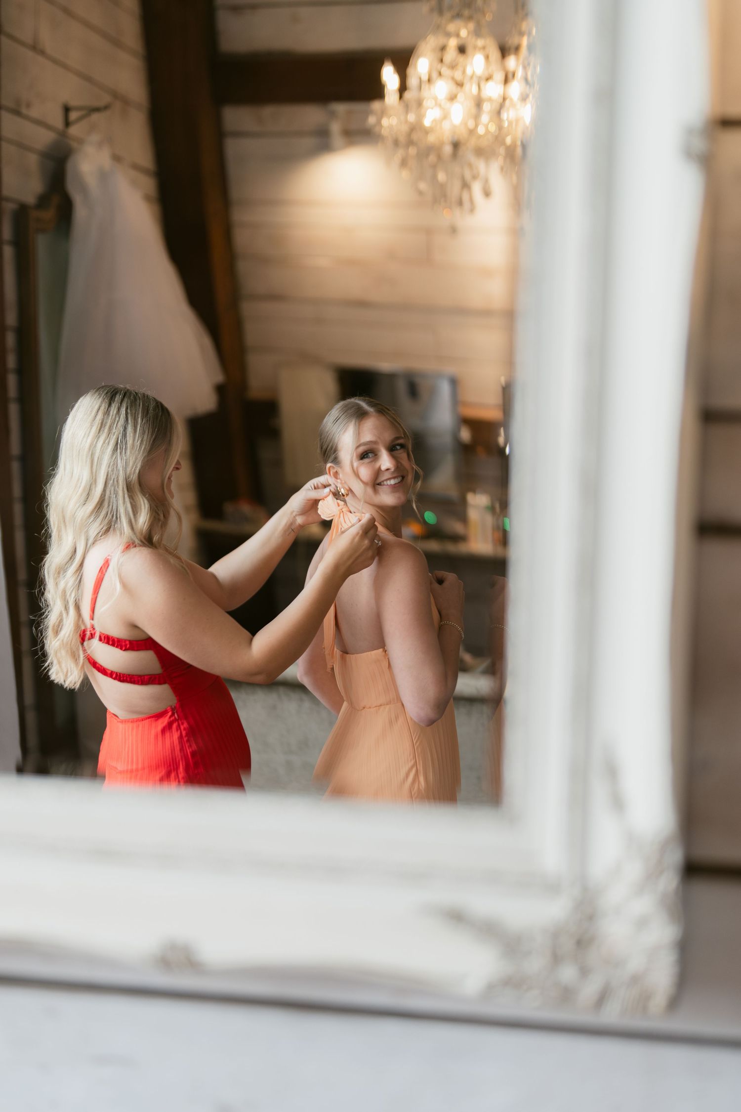 A bridesmaid in red helps a woman in peach dress with earrings before a wedding, reflected in a white mirror.
