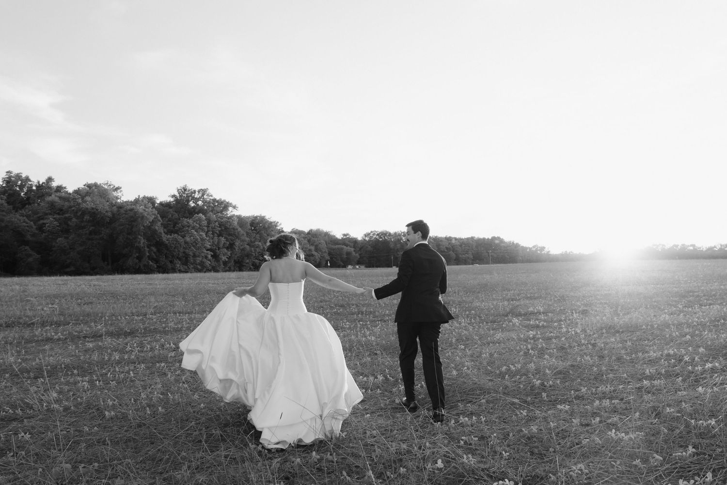 Black and white photo of bride and groom holding hands walking through a sunlit field on their wedding day.