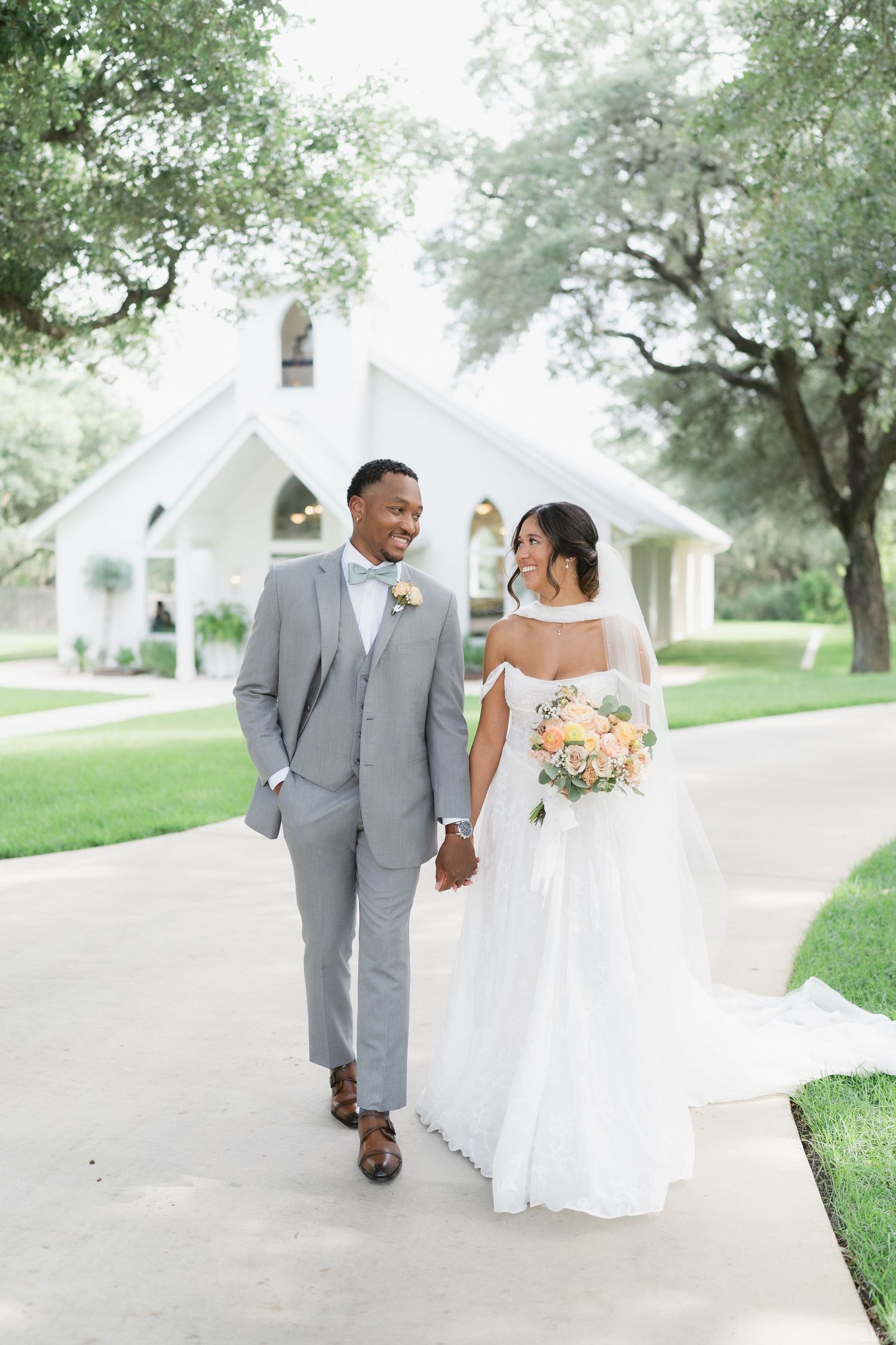 Bride and groom walk hand-in-hand in front of a white chapel, she holds a peach bouquet, he wears a gray suit.