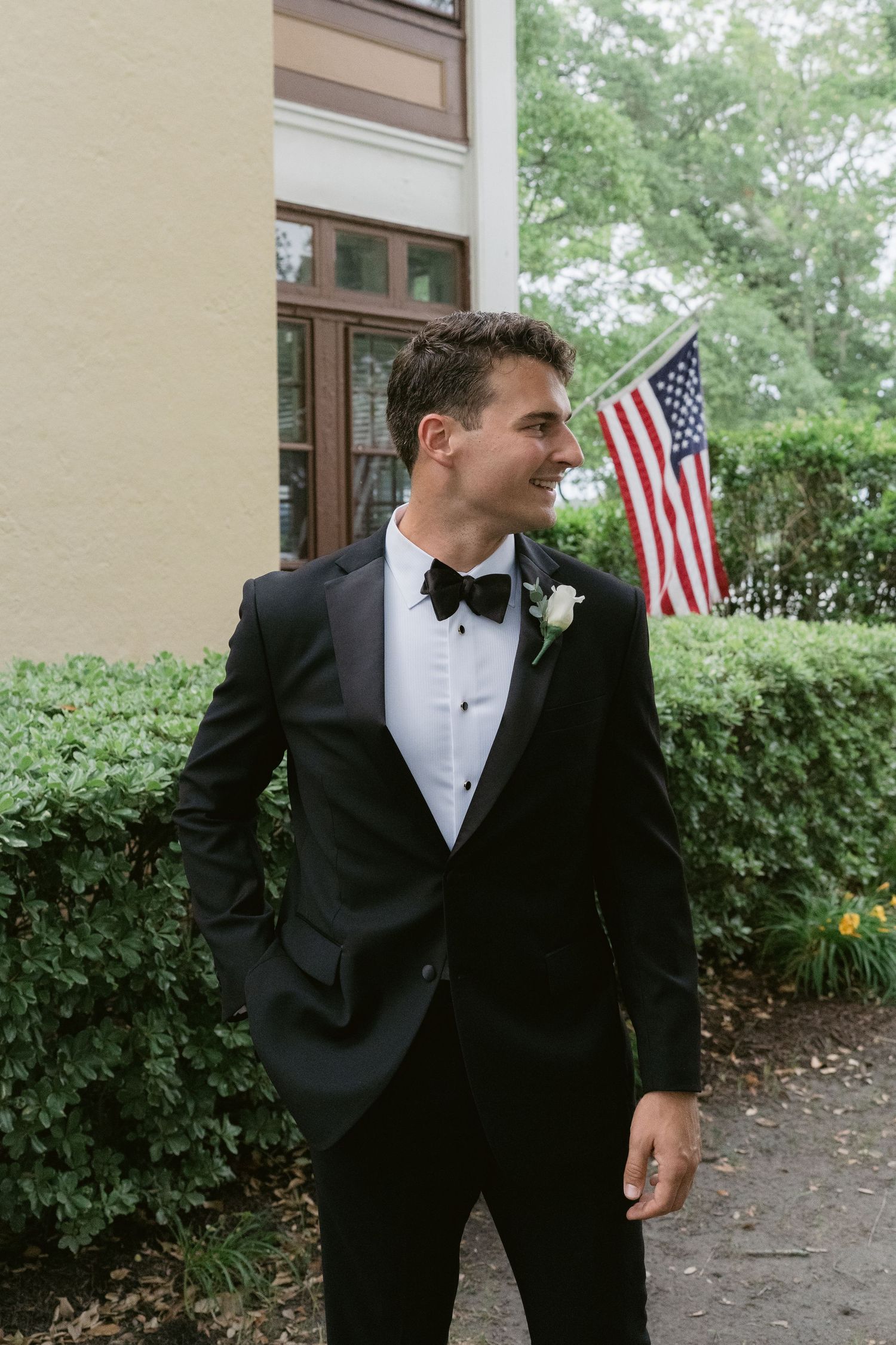 Smiling groom in black tuxedo with bow tie and boutonniere stands outside near an American flag and green hedges.