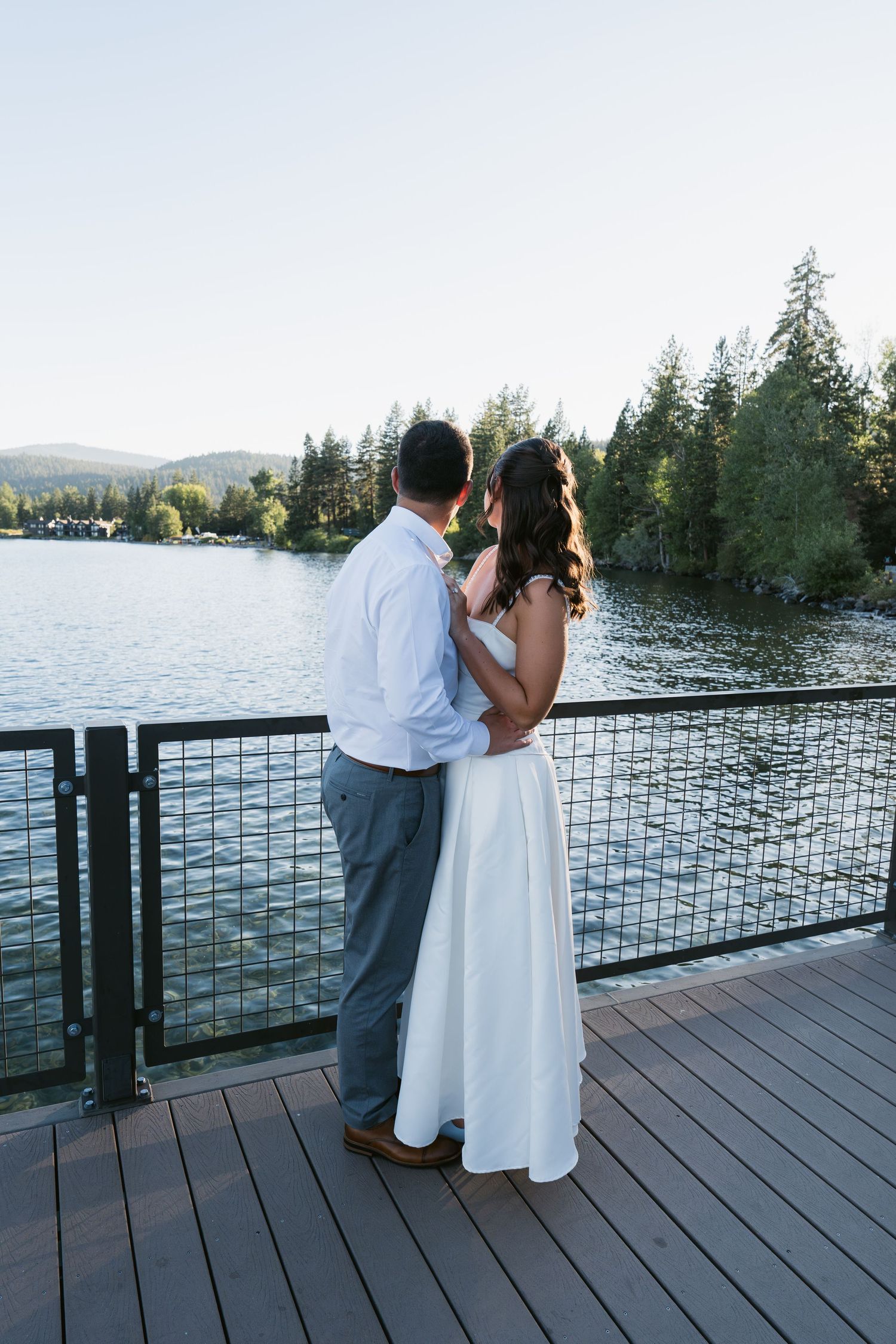 Bride and groom embrace on a wooden deck overlooking a serene lake surrounded by evergreen trees.