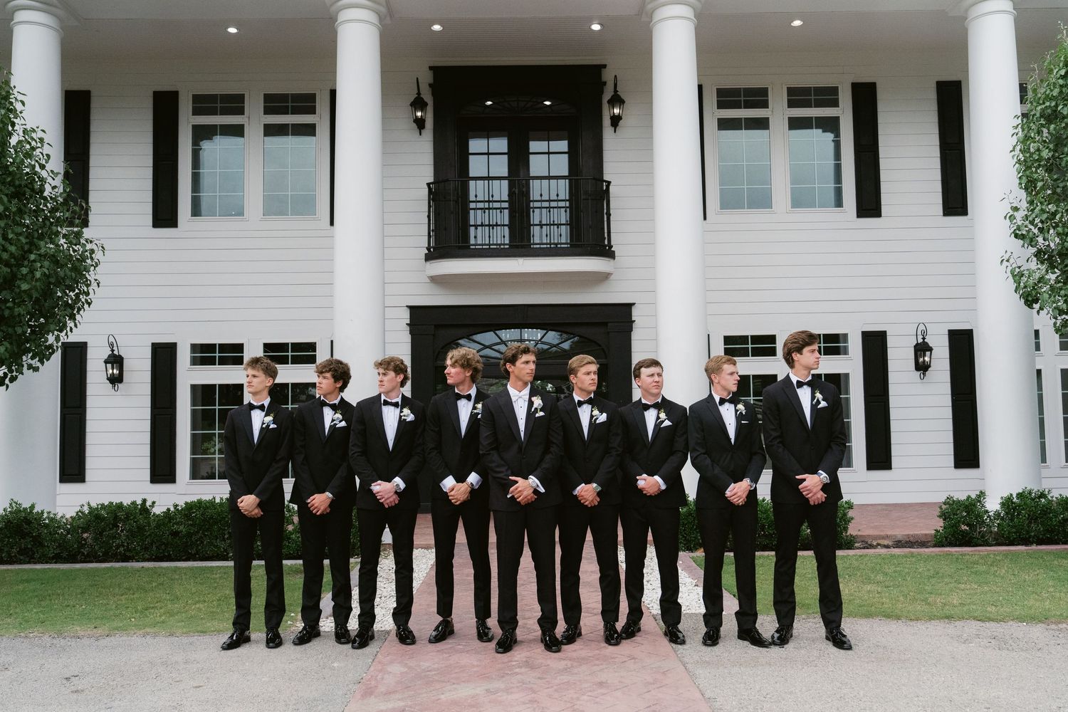 Groomsmen in black tuxedos and bow ties pose in front of a white mansion for a classic wedding photo.