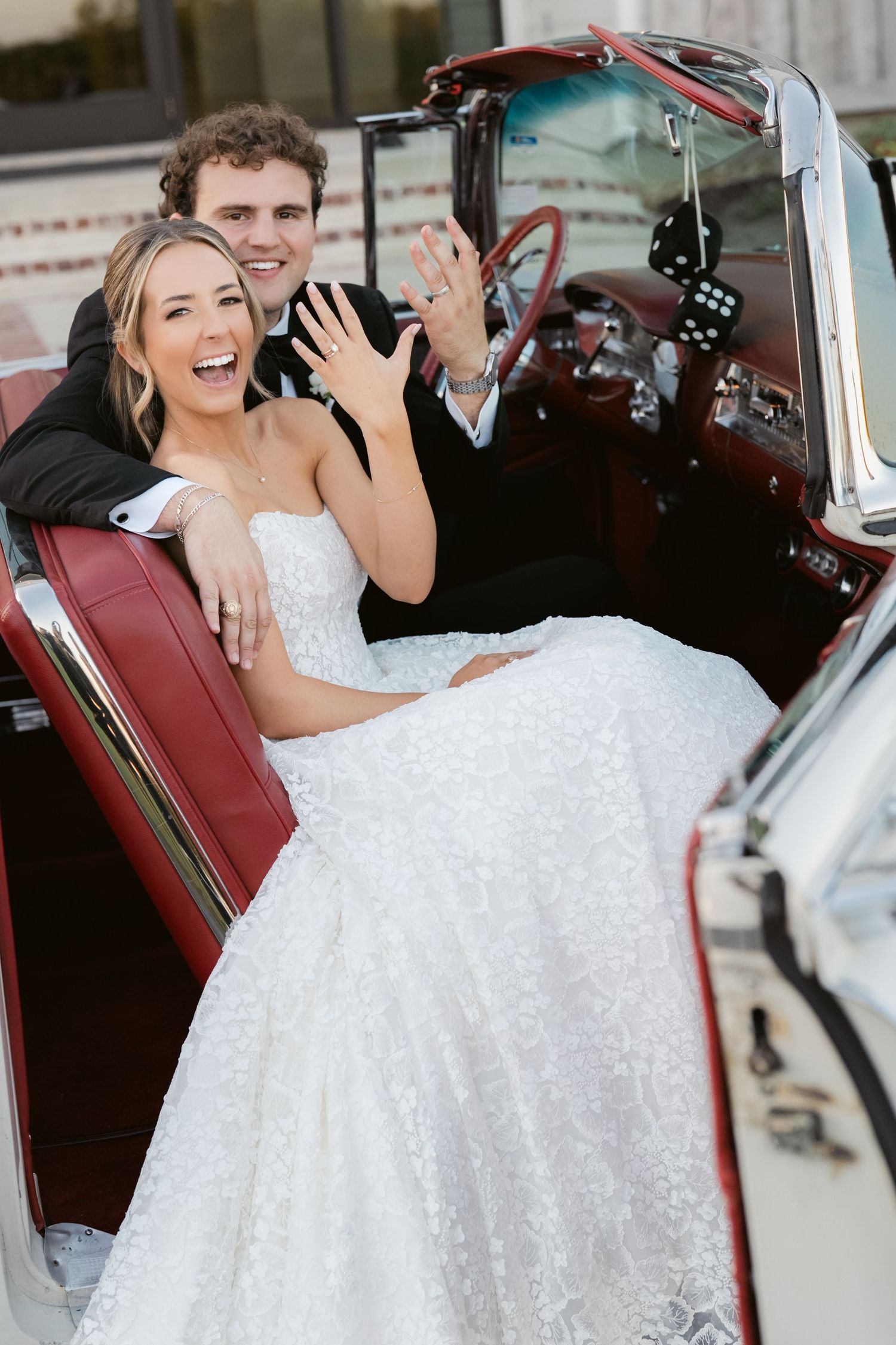 Joyful bride in white lace gown and groom in tuxedo waving from a classic vintage convertible car on their wedding day.