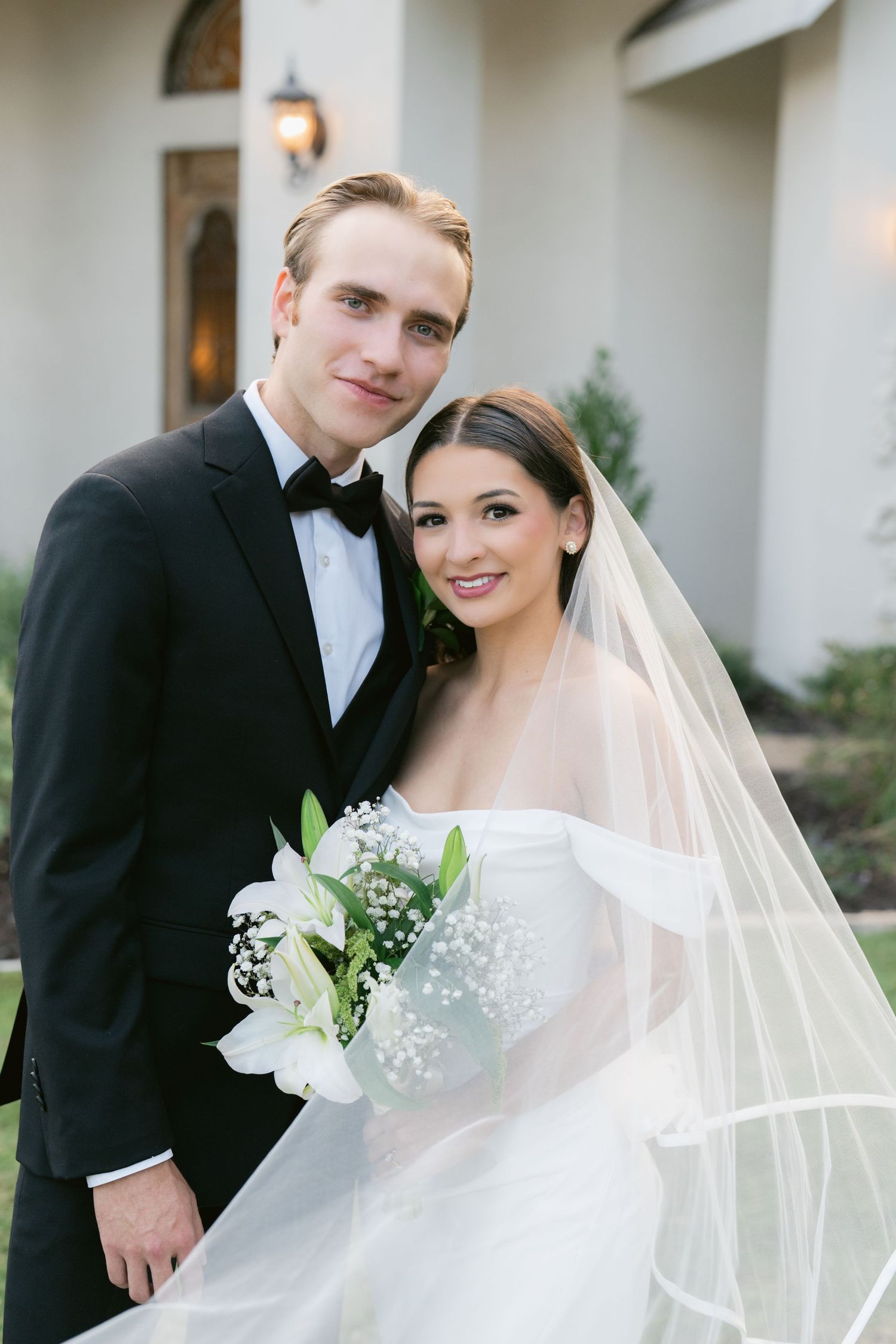 Bride and groom portrait outdoors, she holds white lily bouquet wearing veil, he wears black tuxedo.