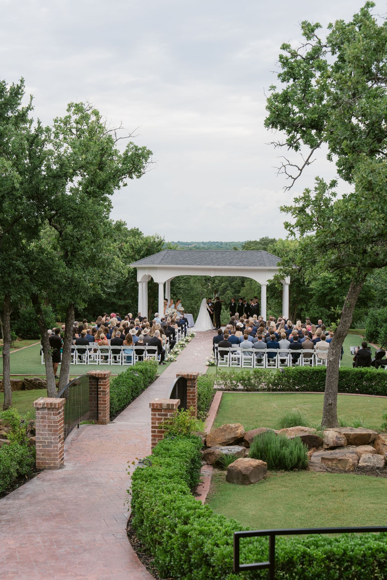 Outdoor wedding ceremony under white gazebo surrounded by lush gardens and seated guests on a cloudy day.