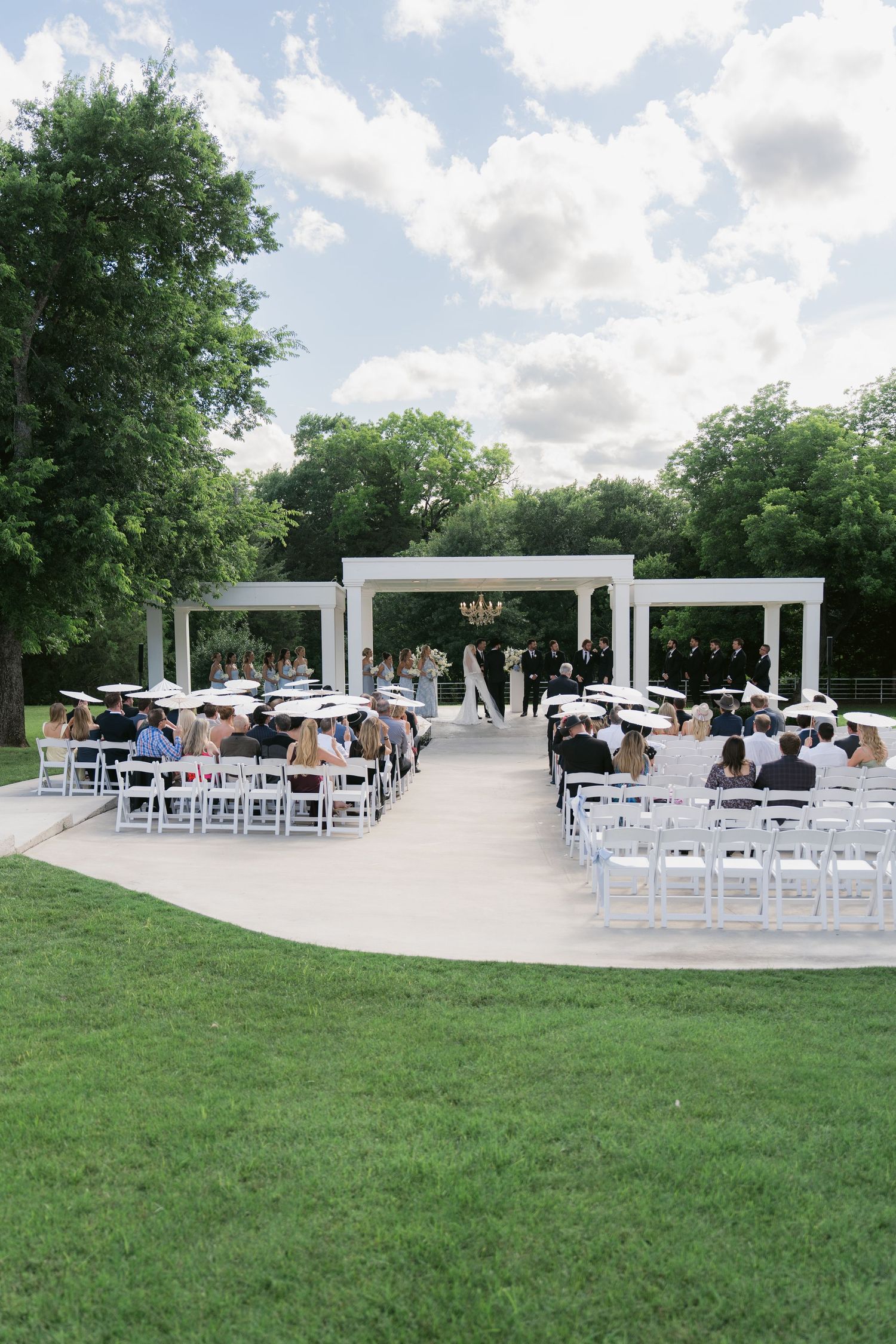 Outdoor wedding ceremony with white chairs, floral arch, chandelier, and lush green trees under a partly cloudy sky.