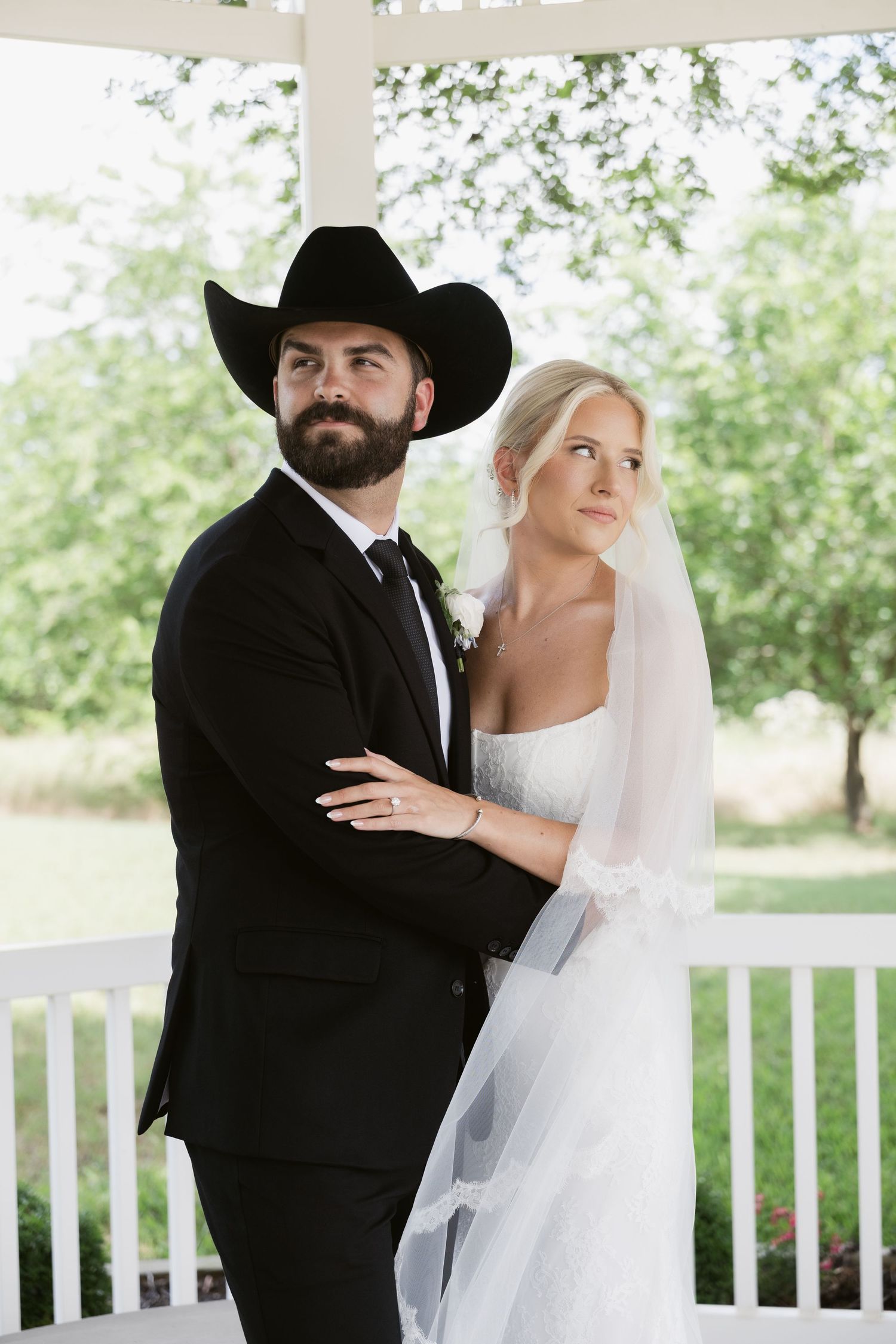 A bearded groom in a black cowboy hat and suit stands with his bride in a white wedding dress and veil on a white porch.