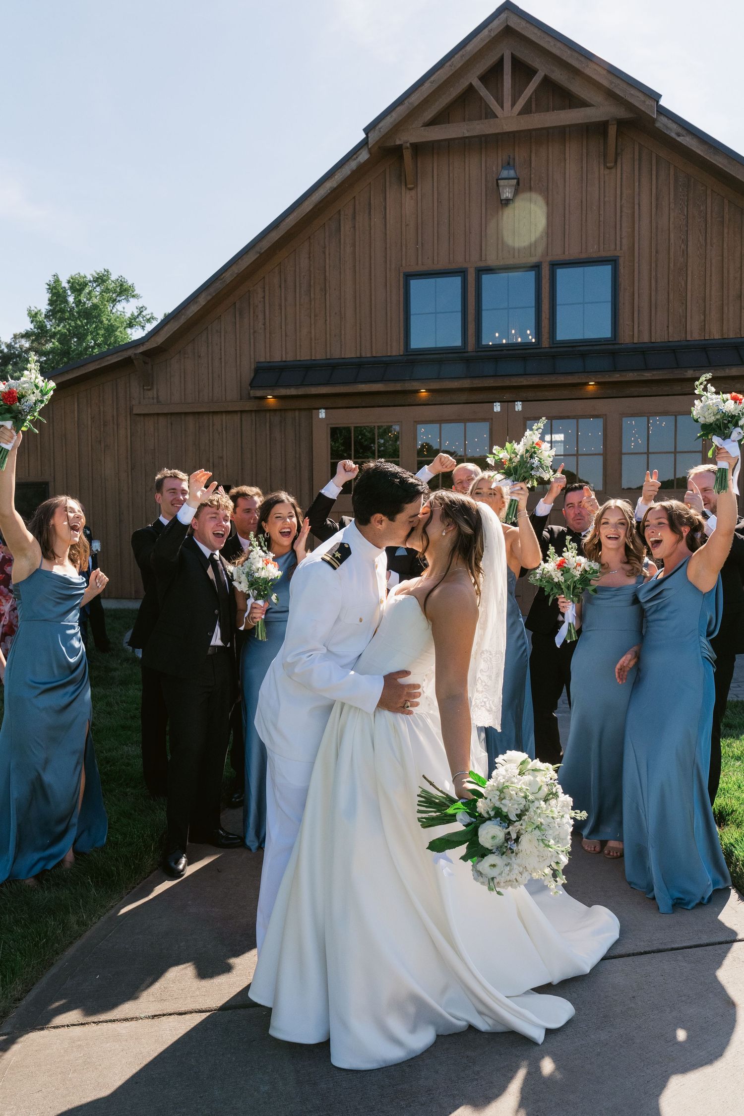 Bride and groom share a kiss surrounded by cheering wedding party in blue gowns and suits outside a rustic barn venue.