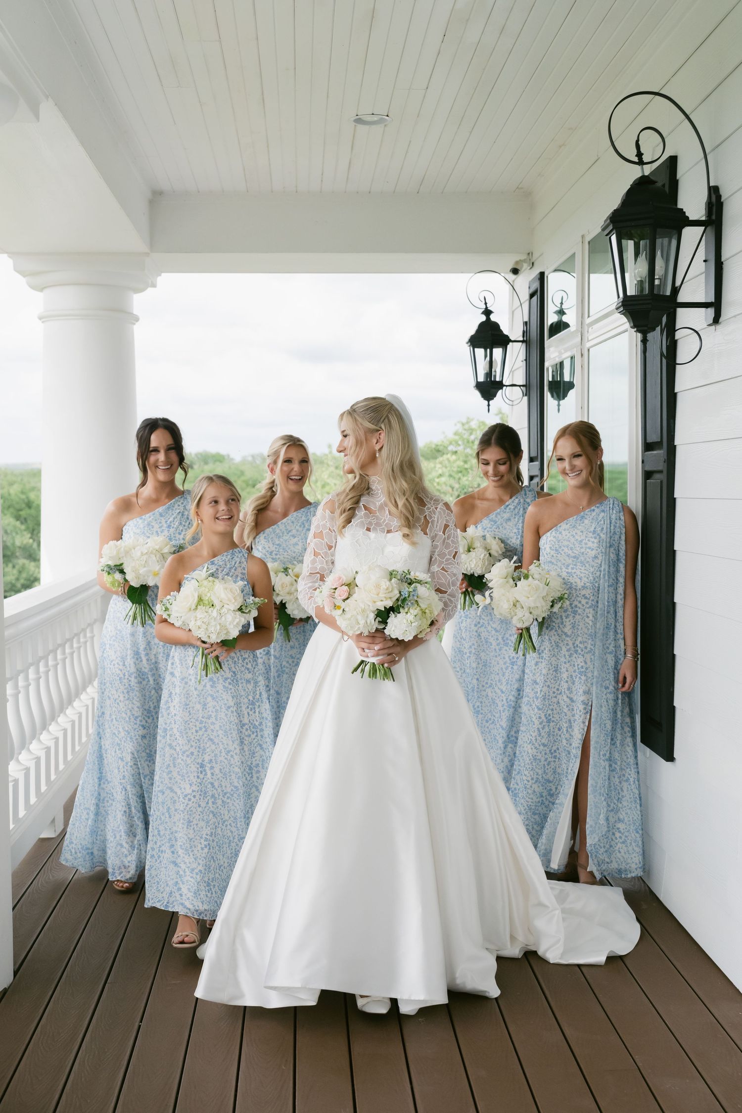 Bride in white gown with bridesmaids in light blue sequin dresses holding white bouquets on a porch.
