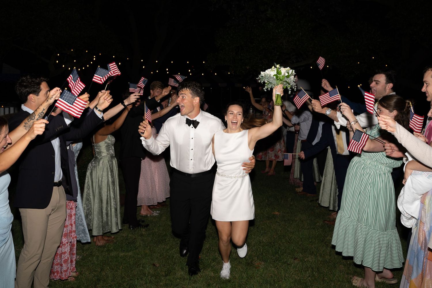 Newlywed couple exits wedding reception through guests waving American flags at night outdoor celebration.