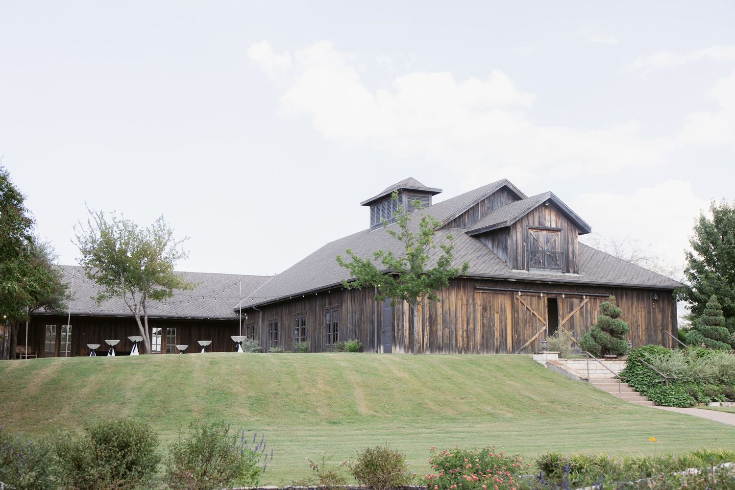 Rustic wooden barn venue with lush green lawn, surrounded by trees under a soft cloudy sky.