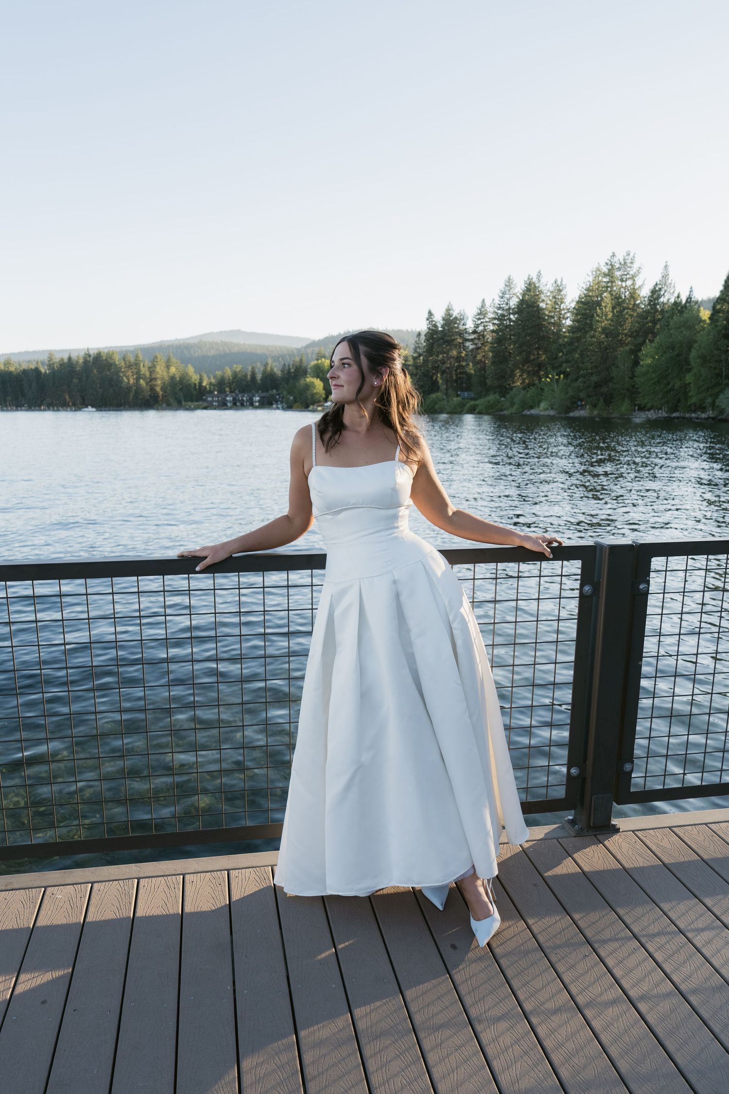 Bride in white gown standing on lakeside deck surrounded by evergreen trees and mountains.