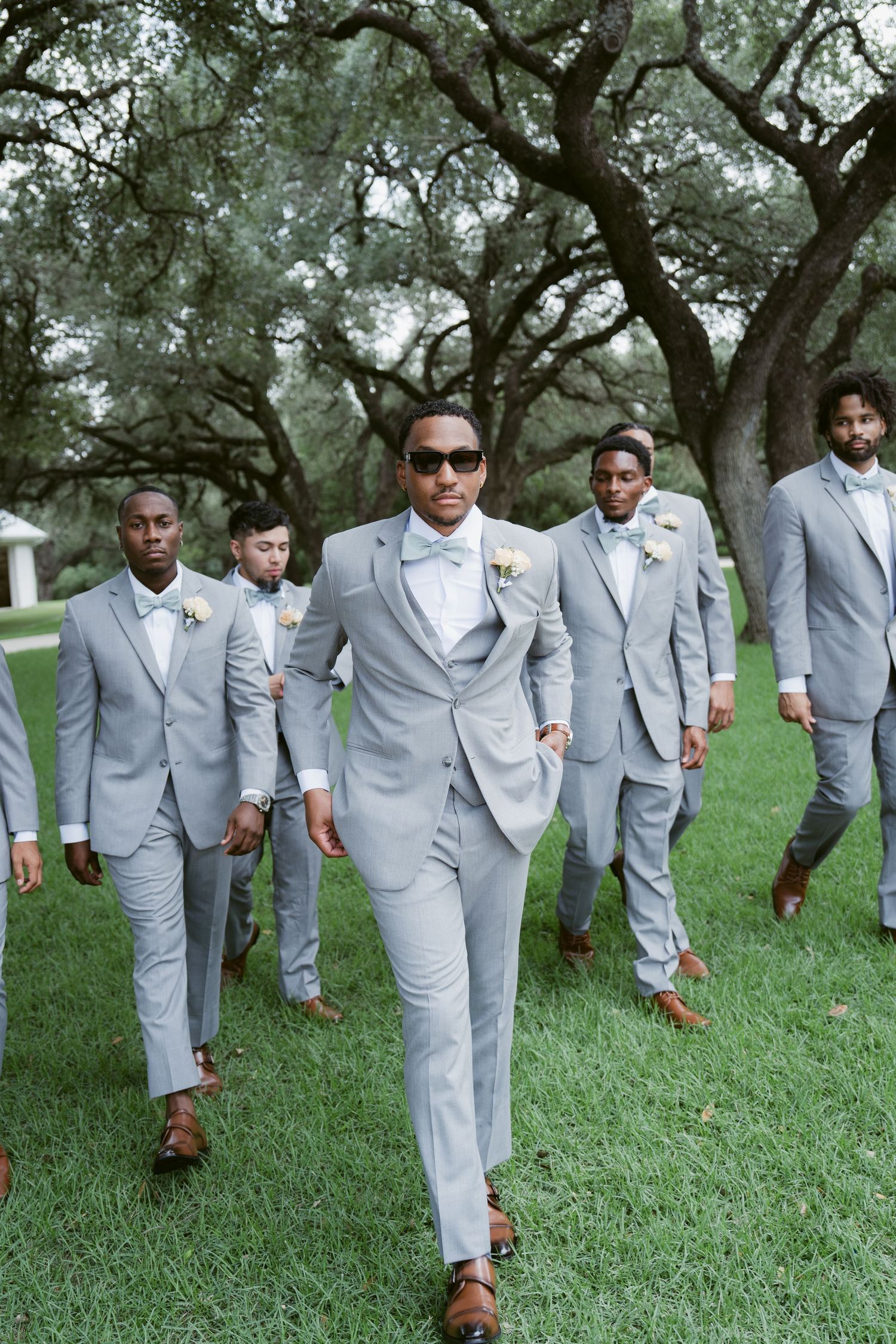 Groom in gray suit and sunglasses leads groomsmen across green lawn under oak trees at outdoor wedding.
