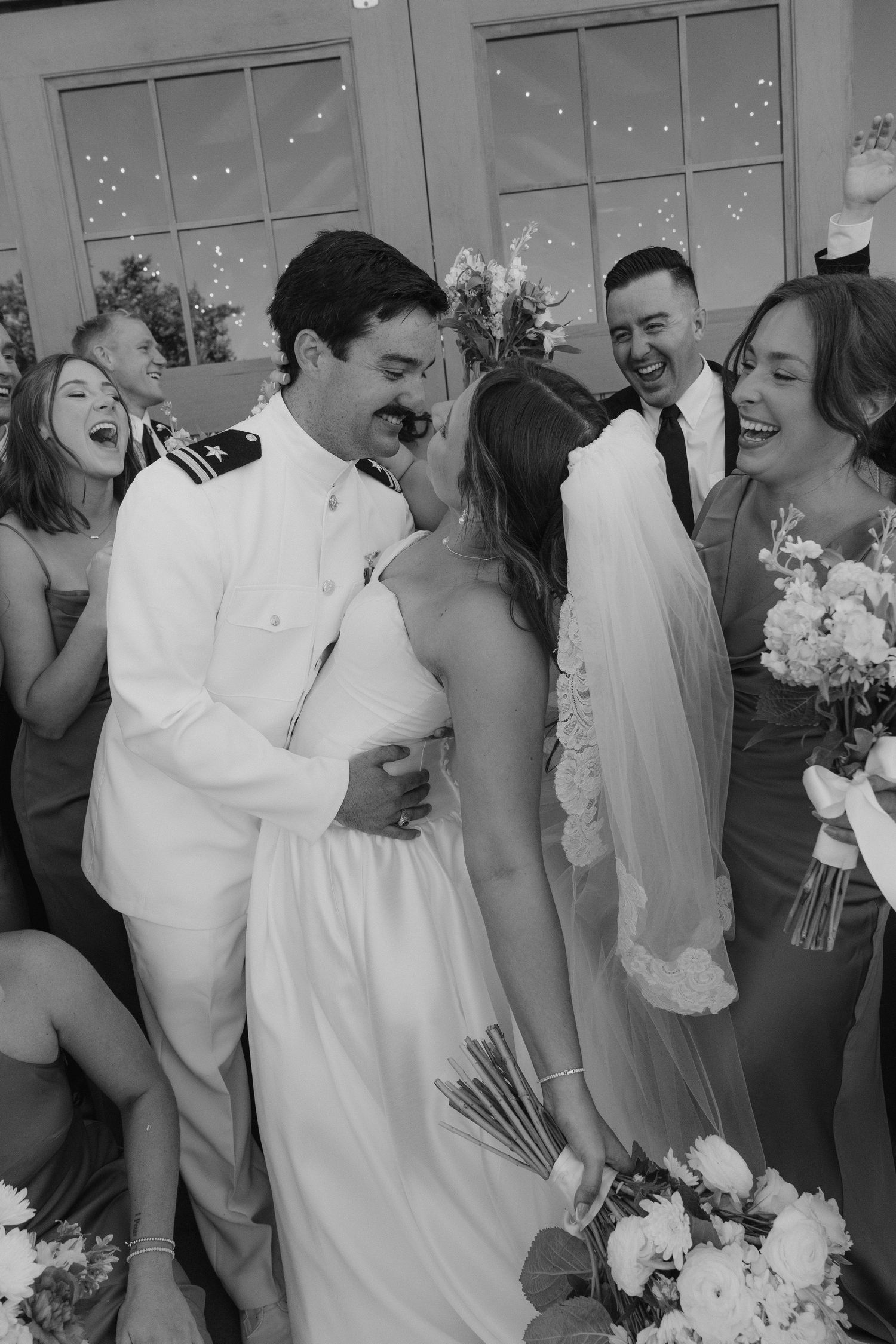 Black and white photo of a joyful bride and groom embracing, surrounded by laughing wedding party members.