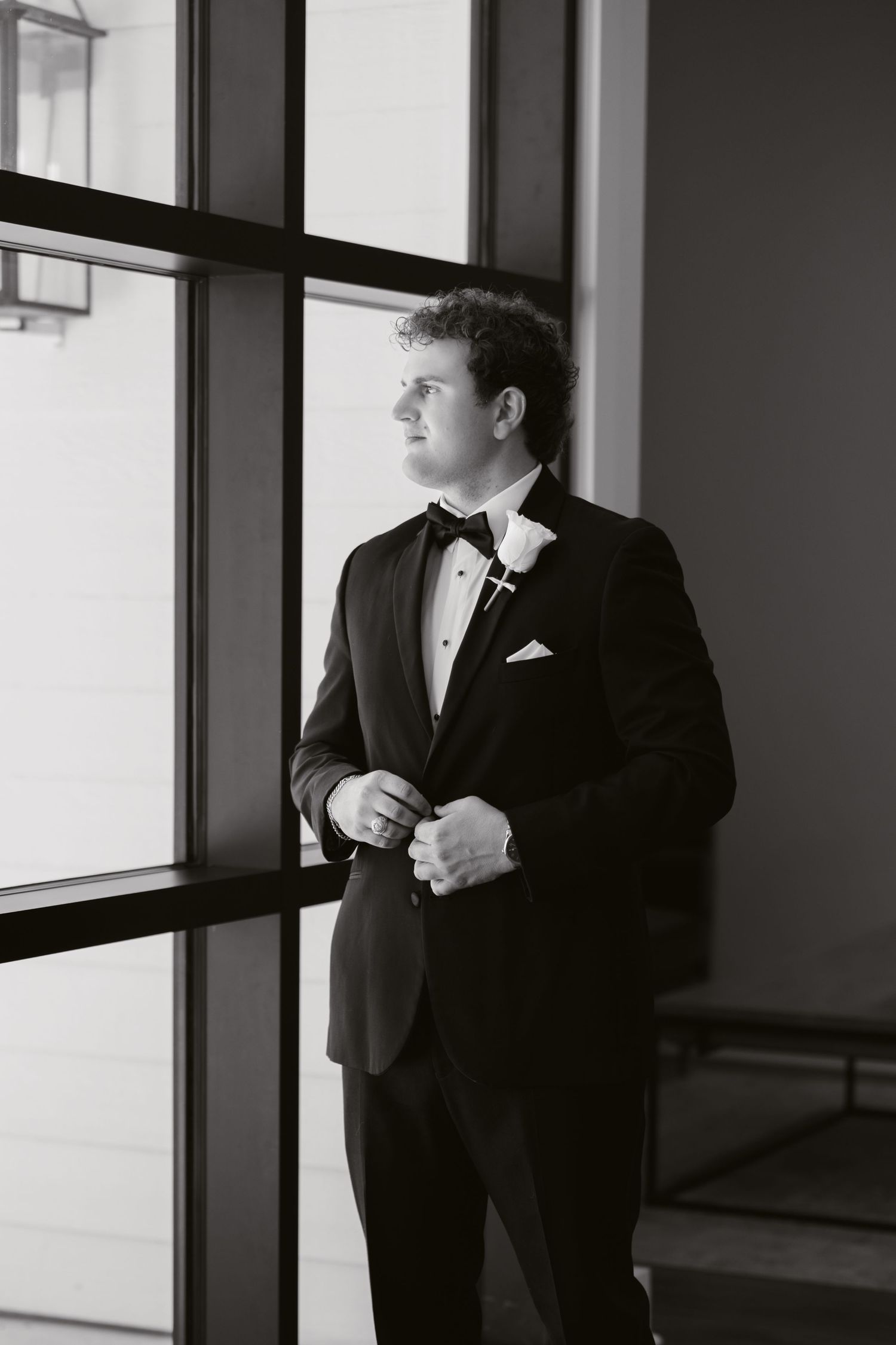 Groom in black tuxedo with bow tie and boutonniere standing by large windows in black and white photo.