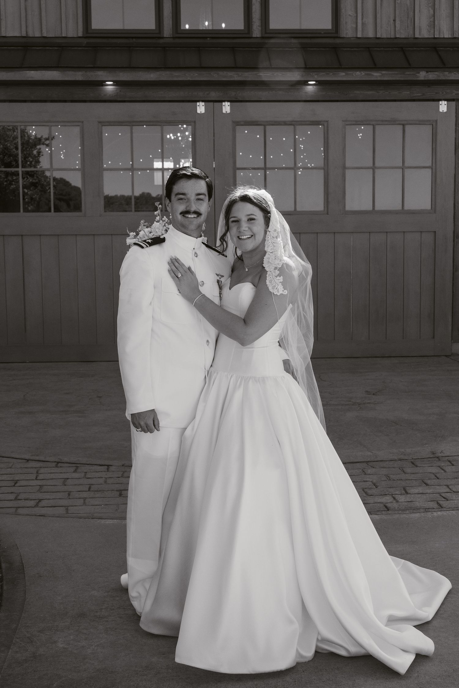 Black and white photo of a smiling bride and groom posing together outside a rustic venue on their wedding day.