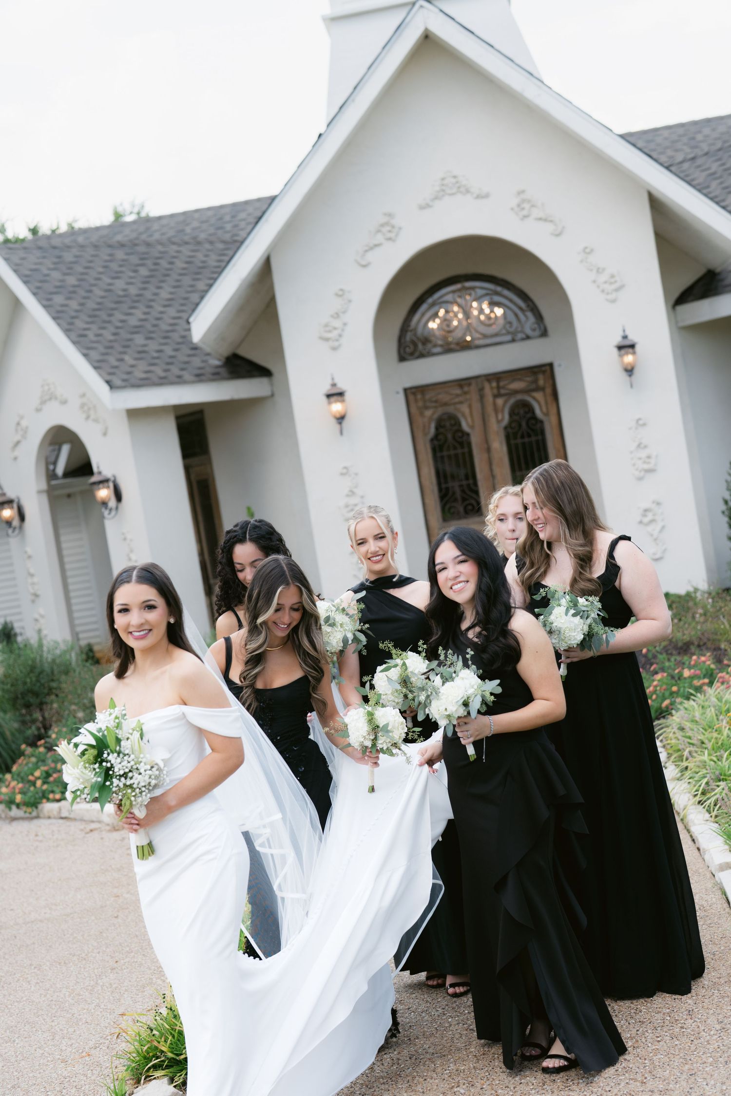 Bride in white gown poses with bridesmaids in black dresses holding white bouquets outside chapel.