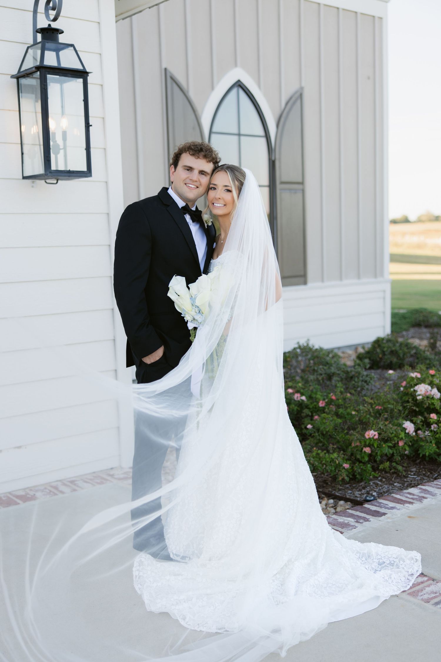 Bride and groom pose outside white chapel, her lace veil flowing in the breeze.