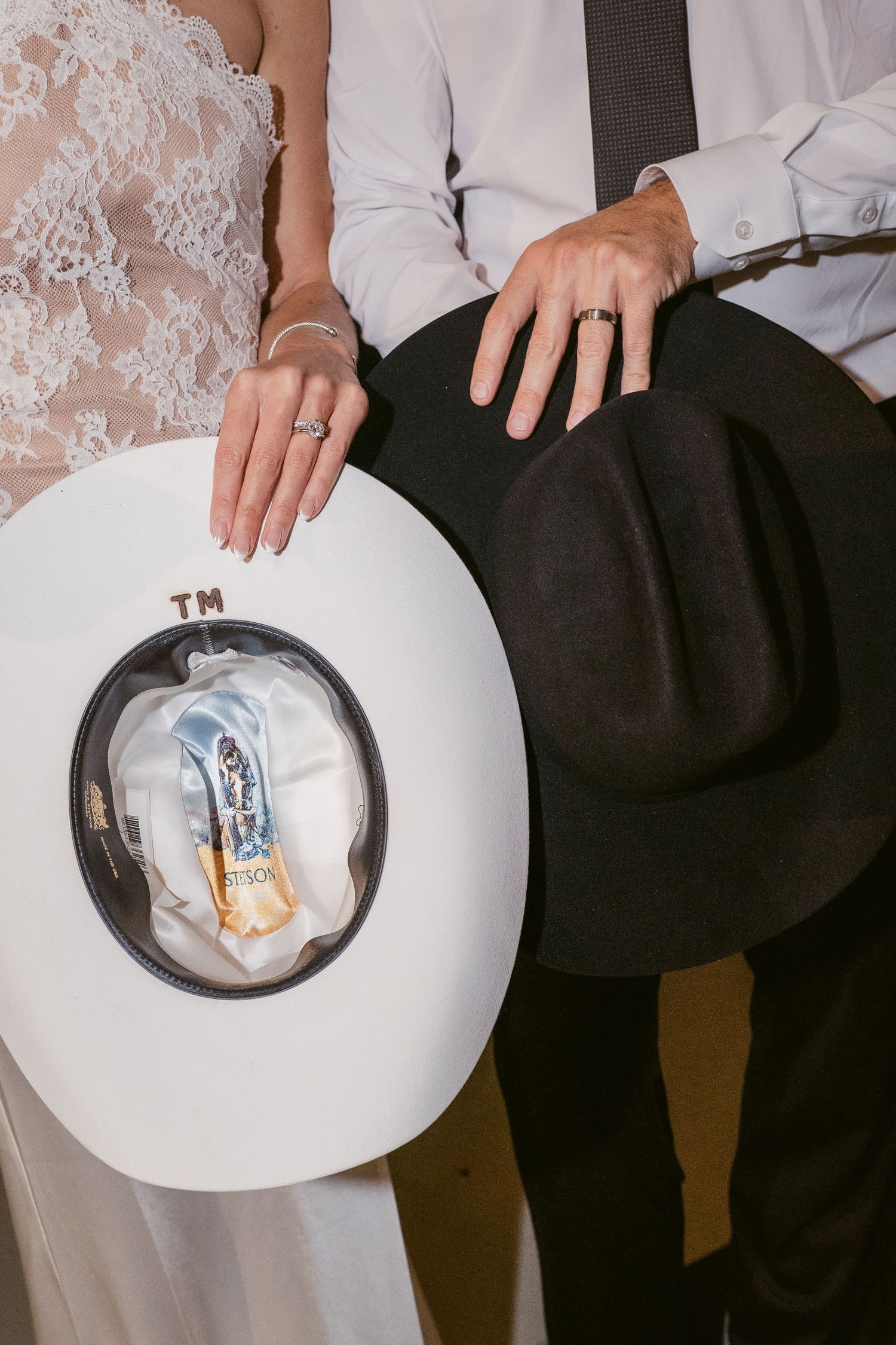 Close-up of bride and groom hands with wedding rings holding white and black cowboy hats with TM initials.