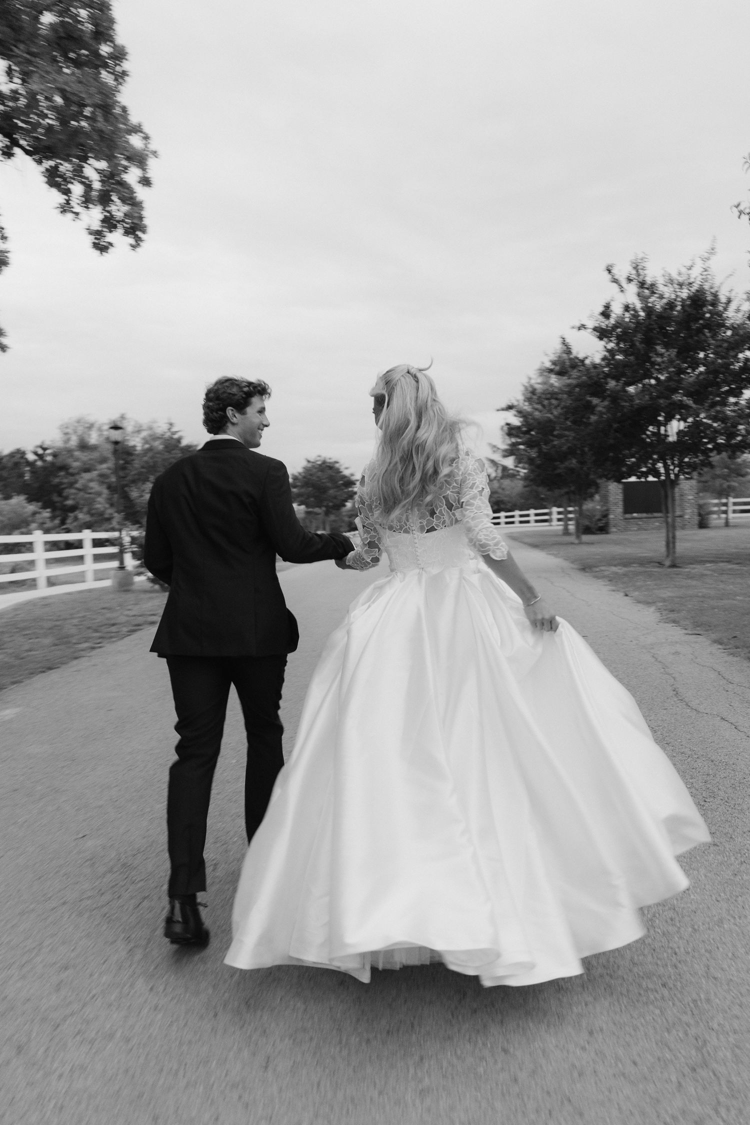 Black and white photo of a bride and groom walking hand in hand down a path on their wedding day.