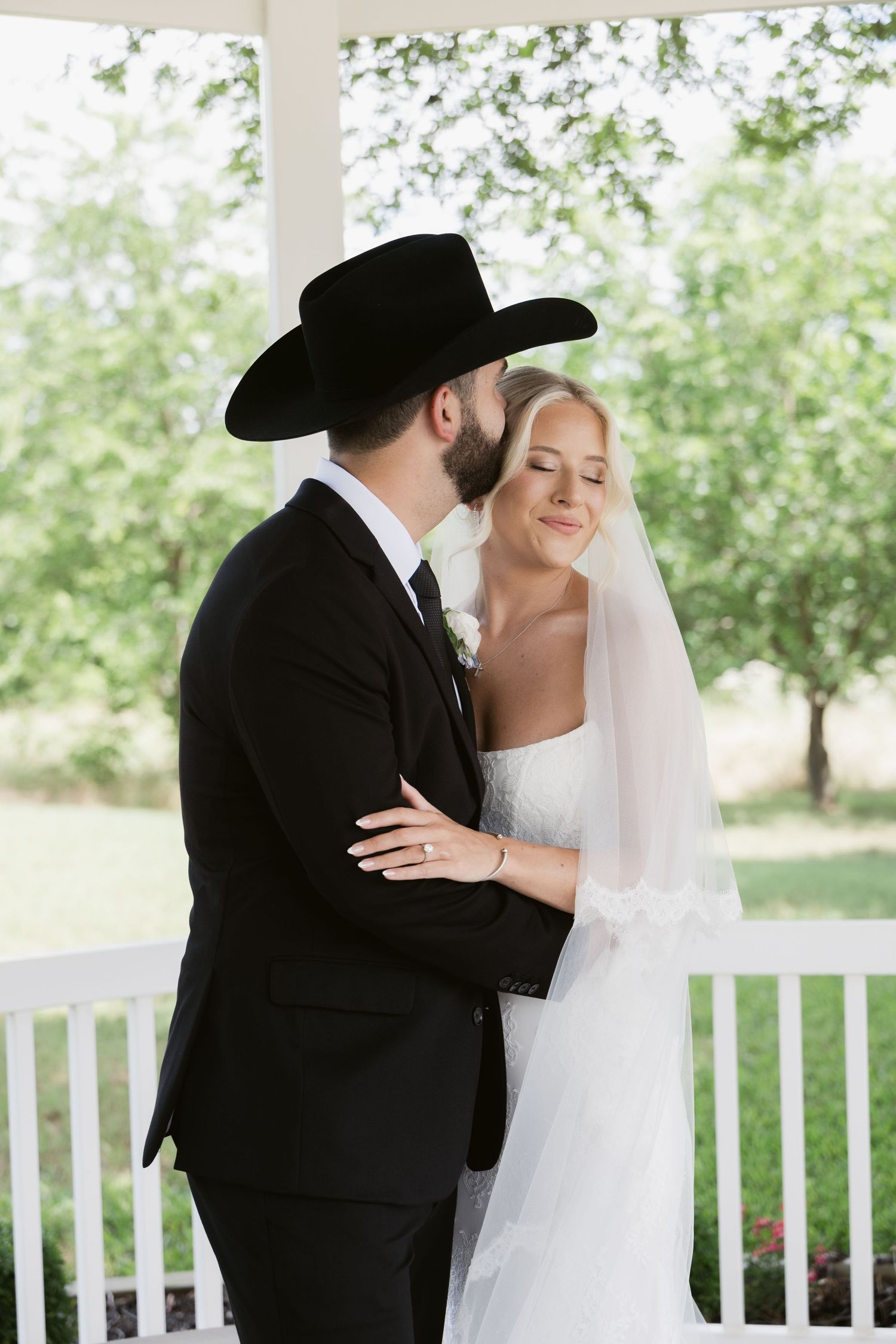 Bride and groom embracing on white porch, groom wearing black cowboy hat and suit, bride in lace wedding dress.