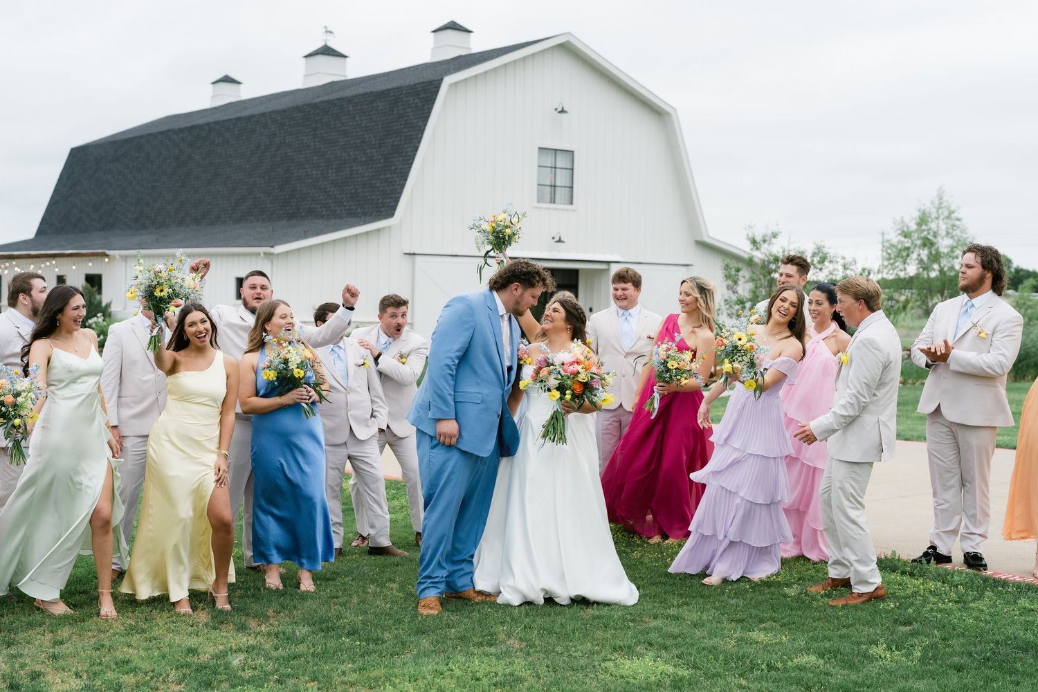 Wedding party in colorful dresses poses with bride and groom kissing in front of a white barn.