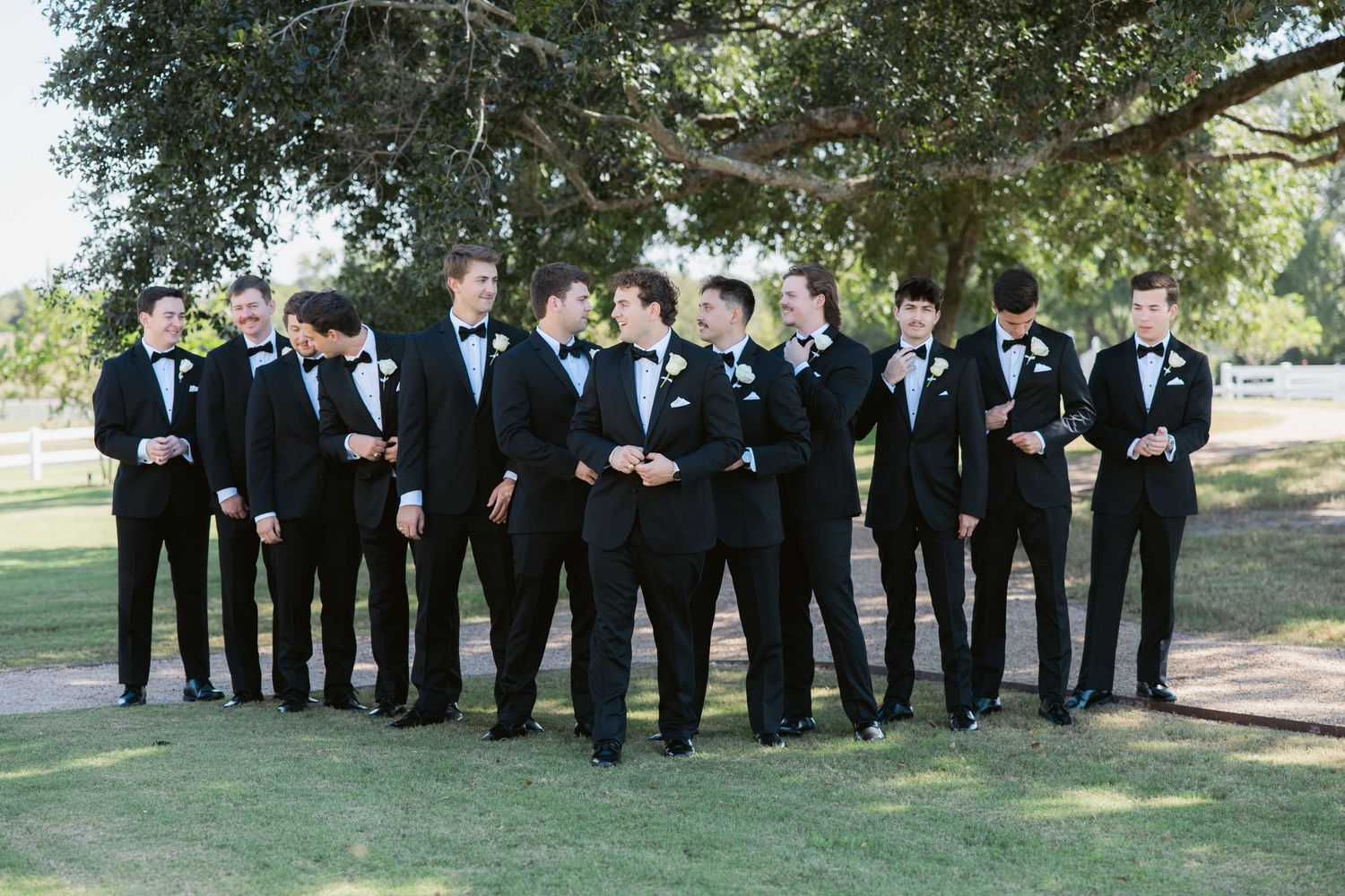 Groom and groomsmen in matching black tuxedos with bow ties gathered outdoors on green lawn for wedding party photo.