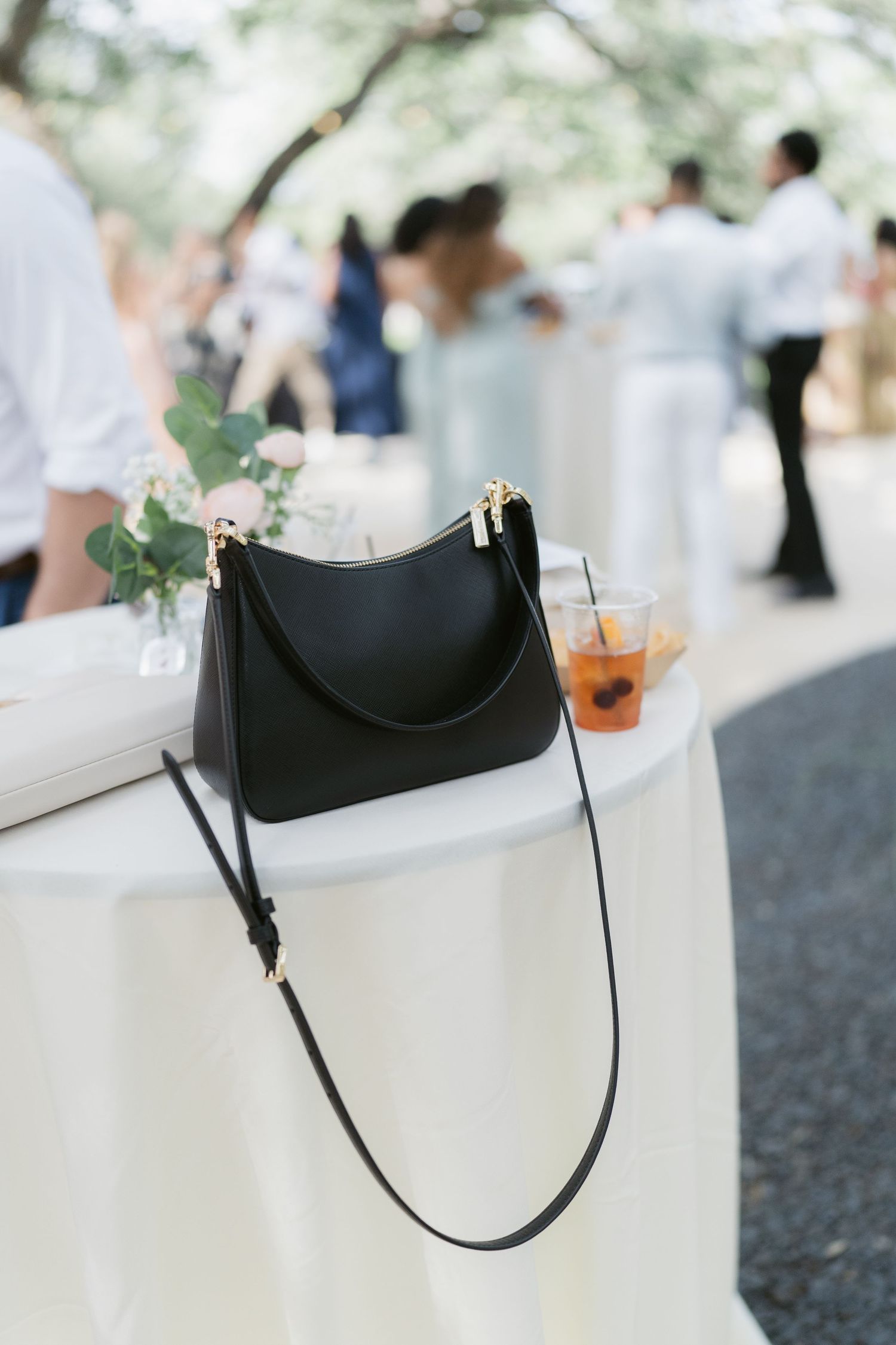 Black leather crossbody bag resting on white tablecloth at outdoor wedding reception with iced drink nearby.