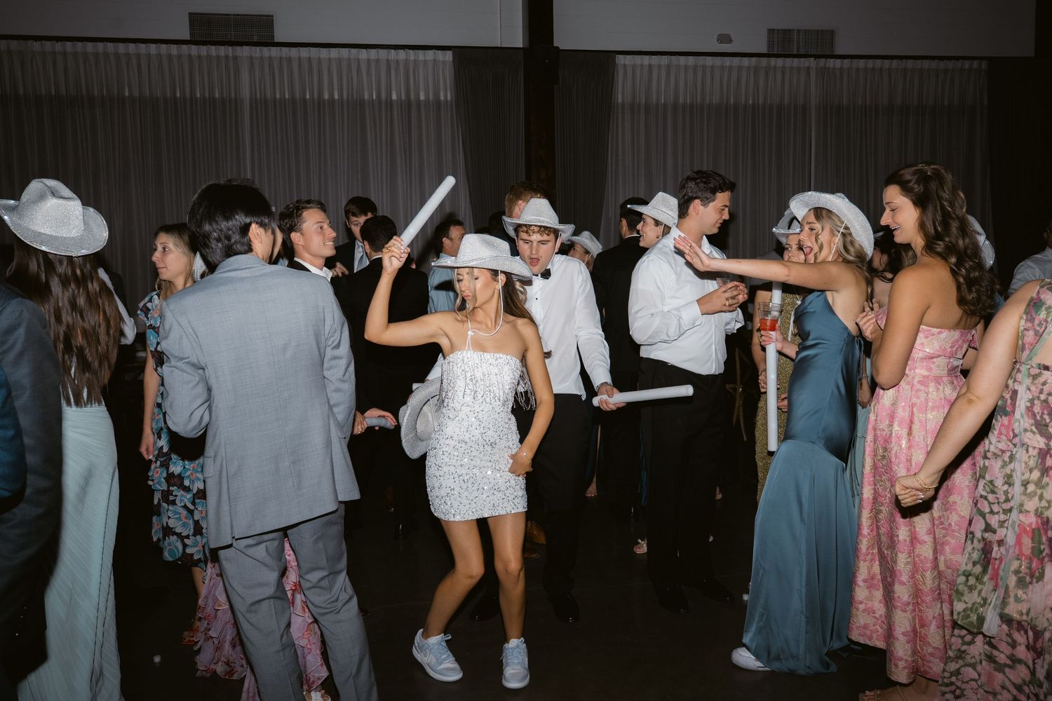 Young adults in formal attire dance at a prom or gala, wearing cowboy hats and holding glow sticks.