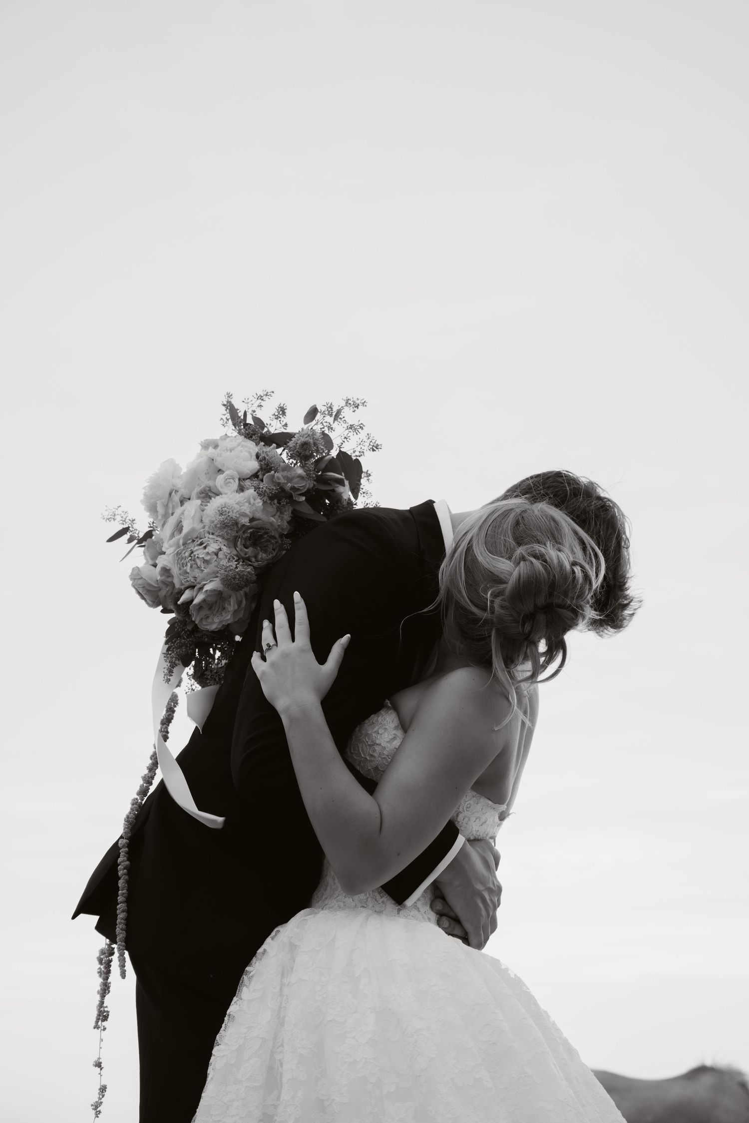 Black and white photo of a bride and groom sharing a kiss, bouquet in hand, on their wedding day.