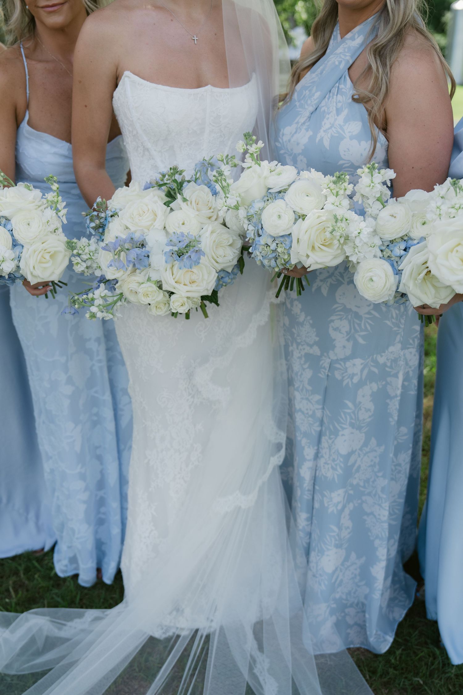 Bride in white lace gown with bridesmaids in light blue dresses holding white and blue floral bouquets.