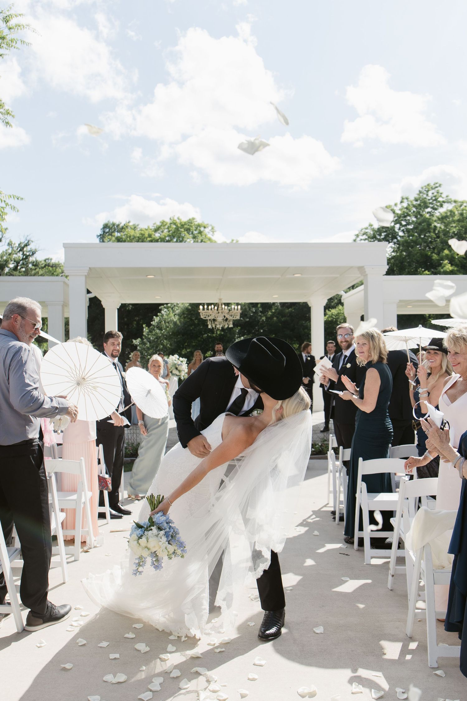 Bride and groom share a romantic dip kiss at outdoor wedding ceremony aisle with guests cheering.