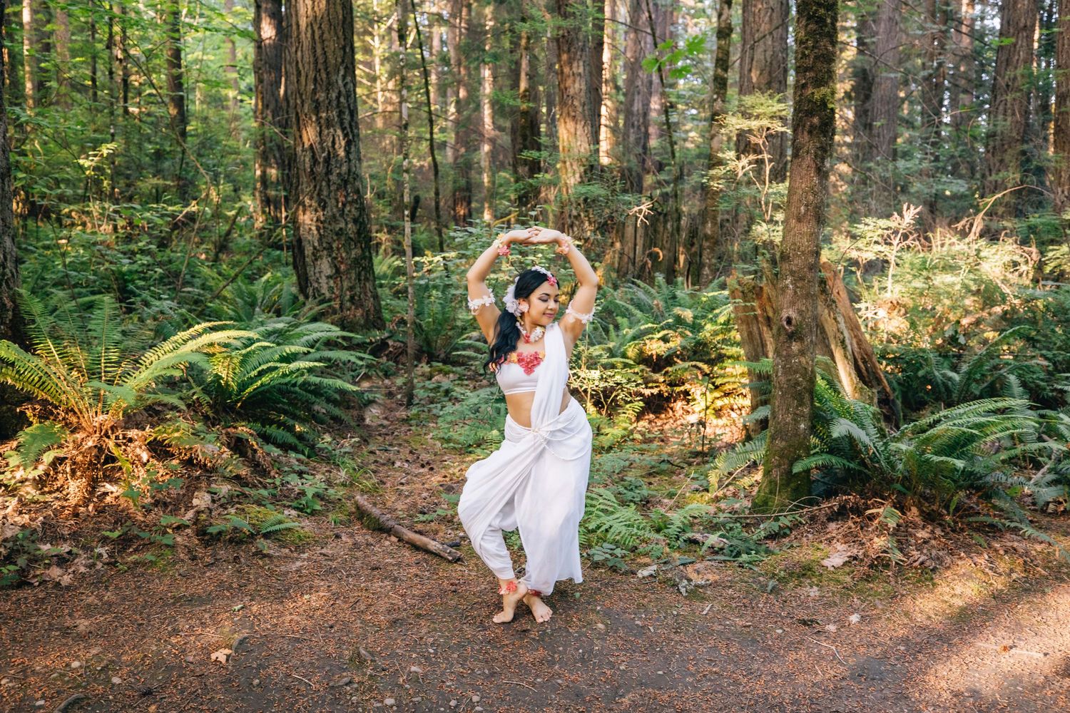 A dancer in white harem pants performs classical Indian dance on a forest path surrounded by ferns and redwood trees.