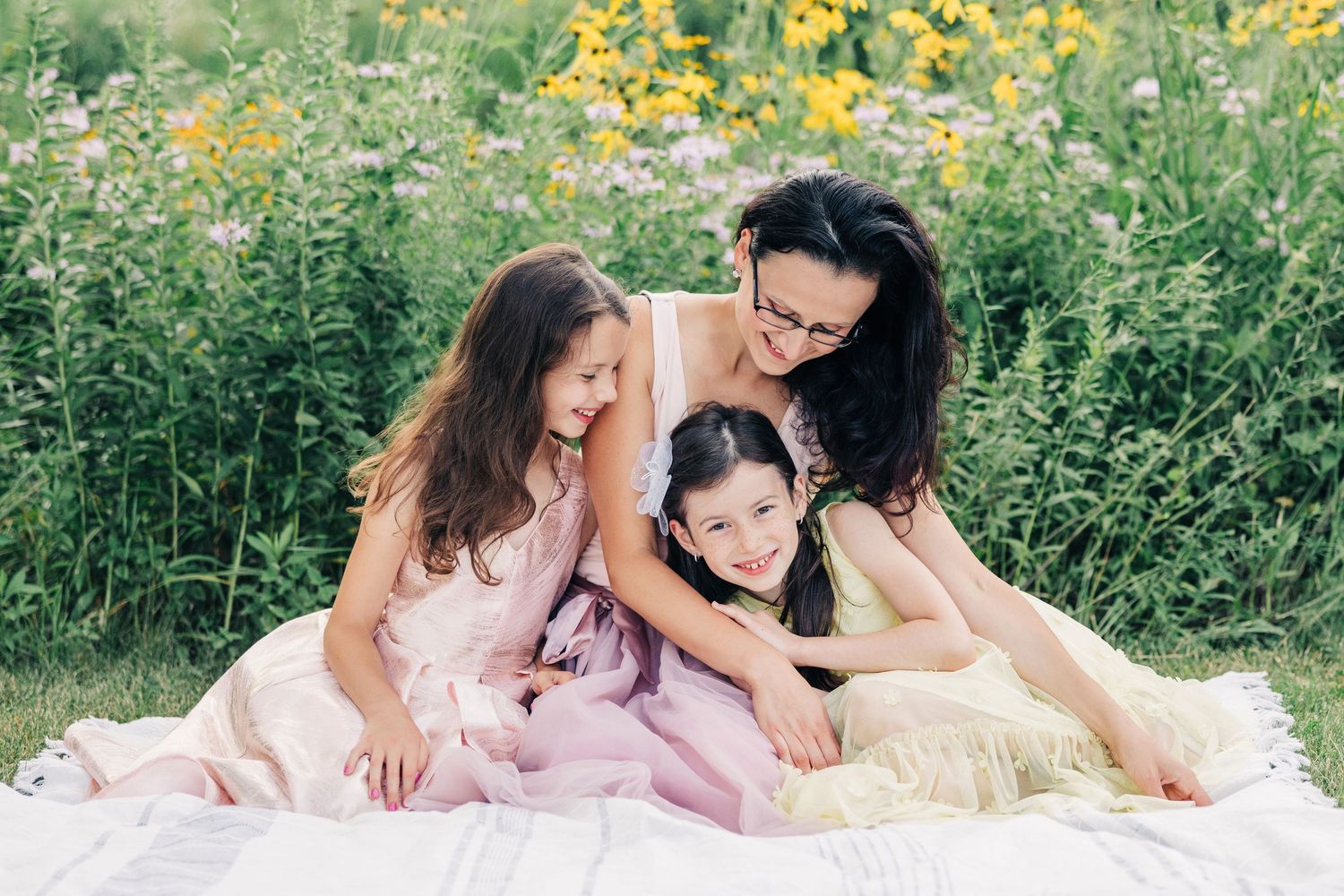 Portrait of a mother and daughters sitting together in field of blooming sunflowers wearing pink dresses.