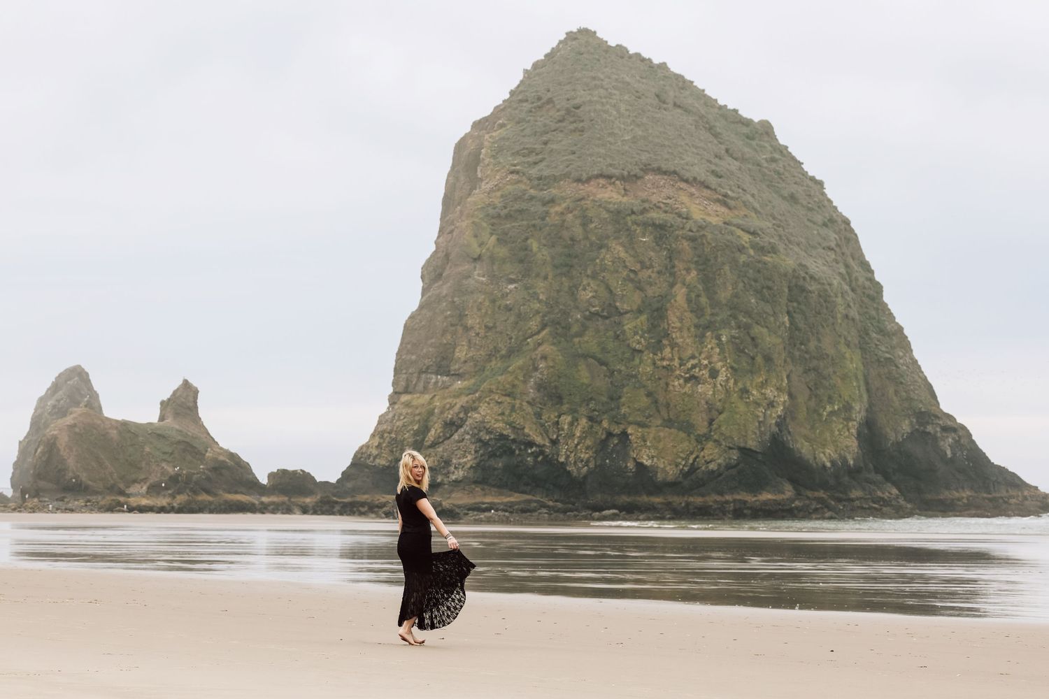 Woman in black dress walking on Cannon Beach with iconic Haystack Rock in the background on an overcast day.