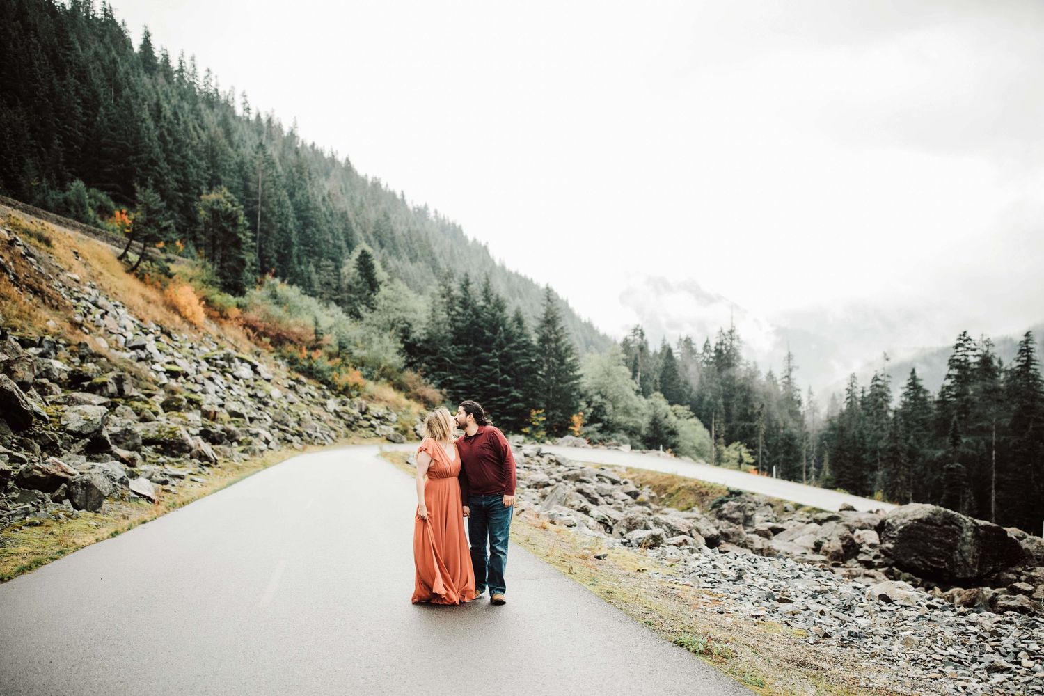 Couple embracing on a misty mountain road surrounded by evergreen forests and rocky terrain.