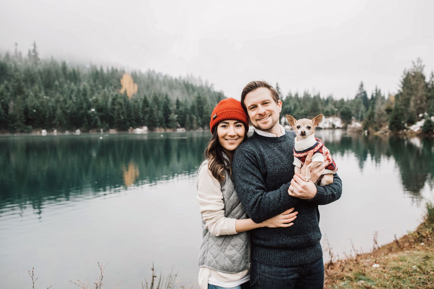 Happy couple holding a small dog by a serene mountain lake with evergreen trees in the background.