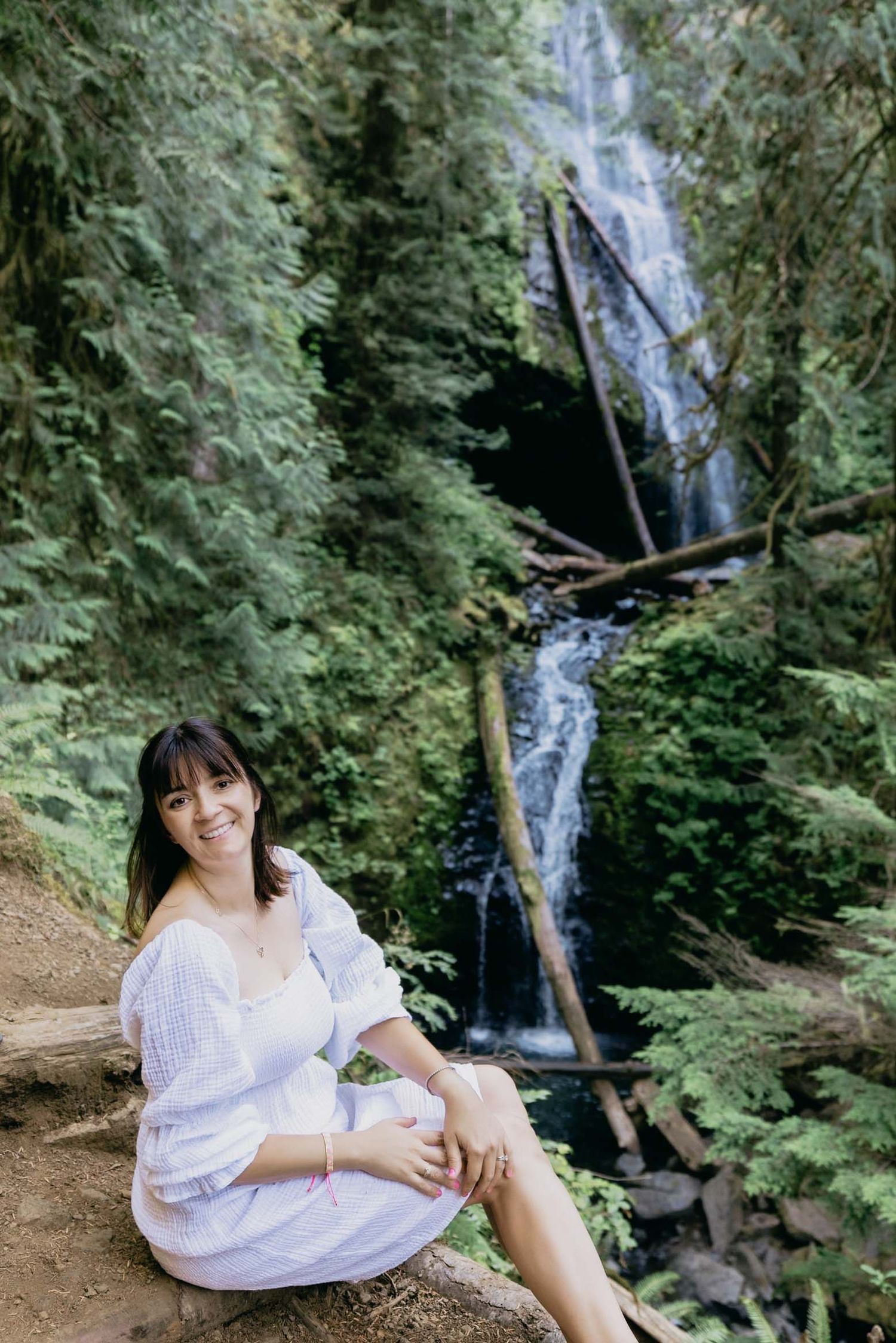 Woman in white dress smiling on rocks before lush green forest waterfall.