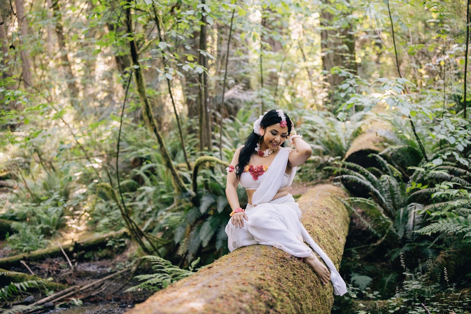 A bride in a white dress sits on a moss-covered log in a lush green forest setting.