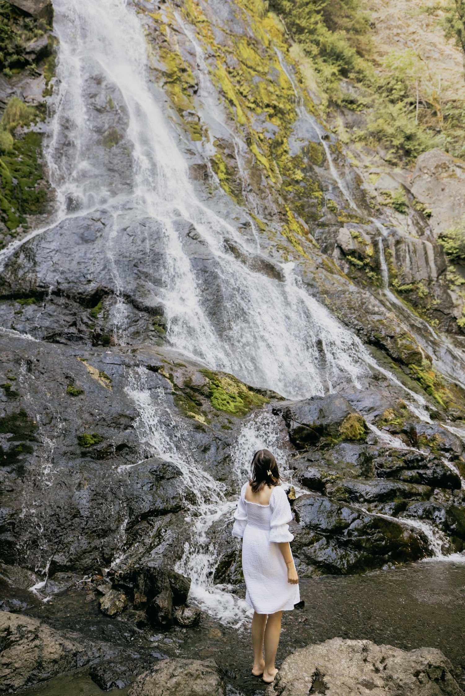 Woman in white off-shoulder dress standing before a cascading waterfall surrounded by mossy rocks.