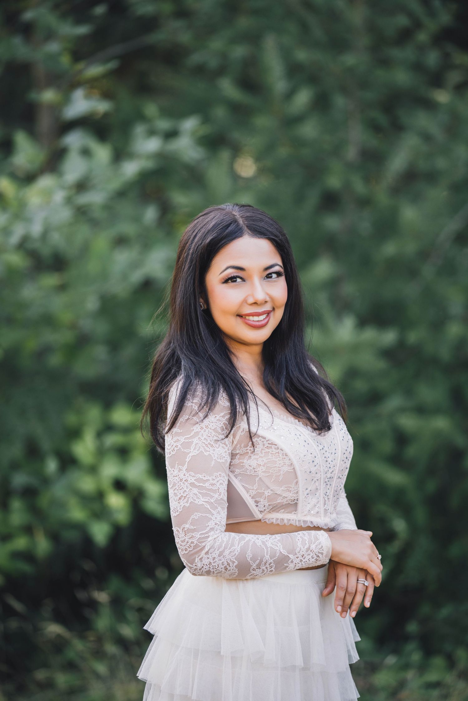 Portrait of a girl in a person in a white lace dress smiles while posing outdoors against a green natural background.