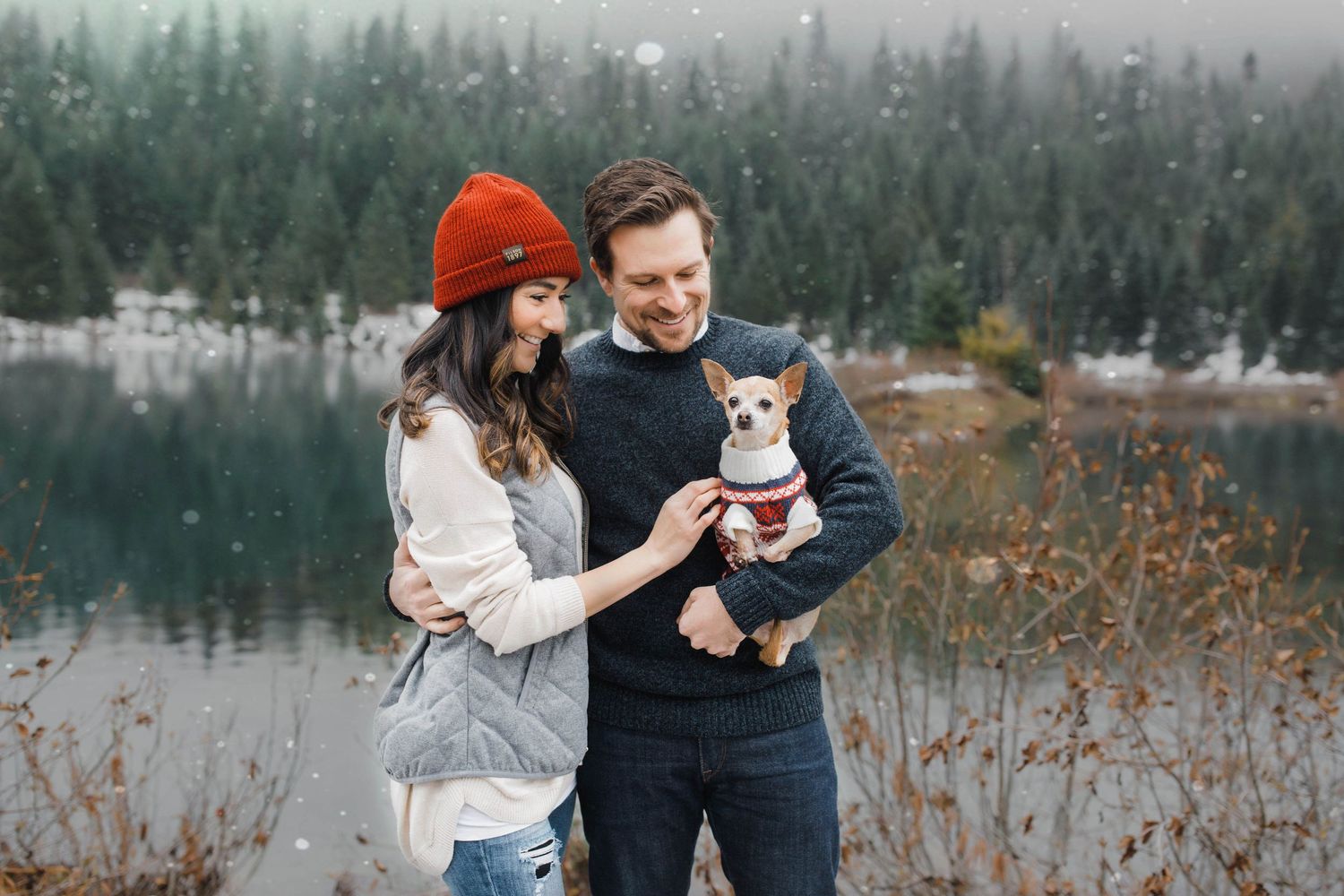 A photo couple stands by a misty lake in winter holding their small white dog while wearing cozy sweaters.